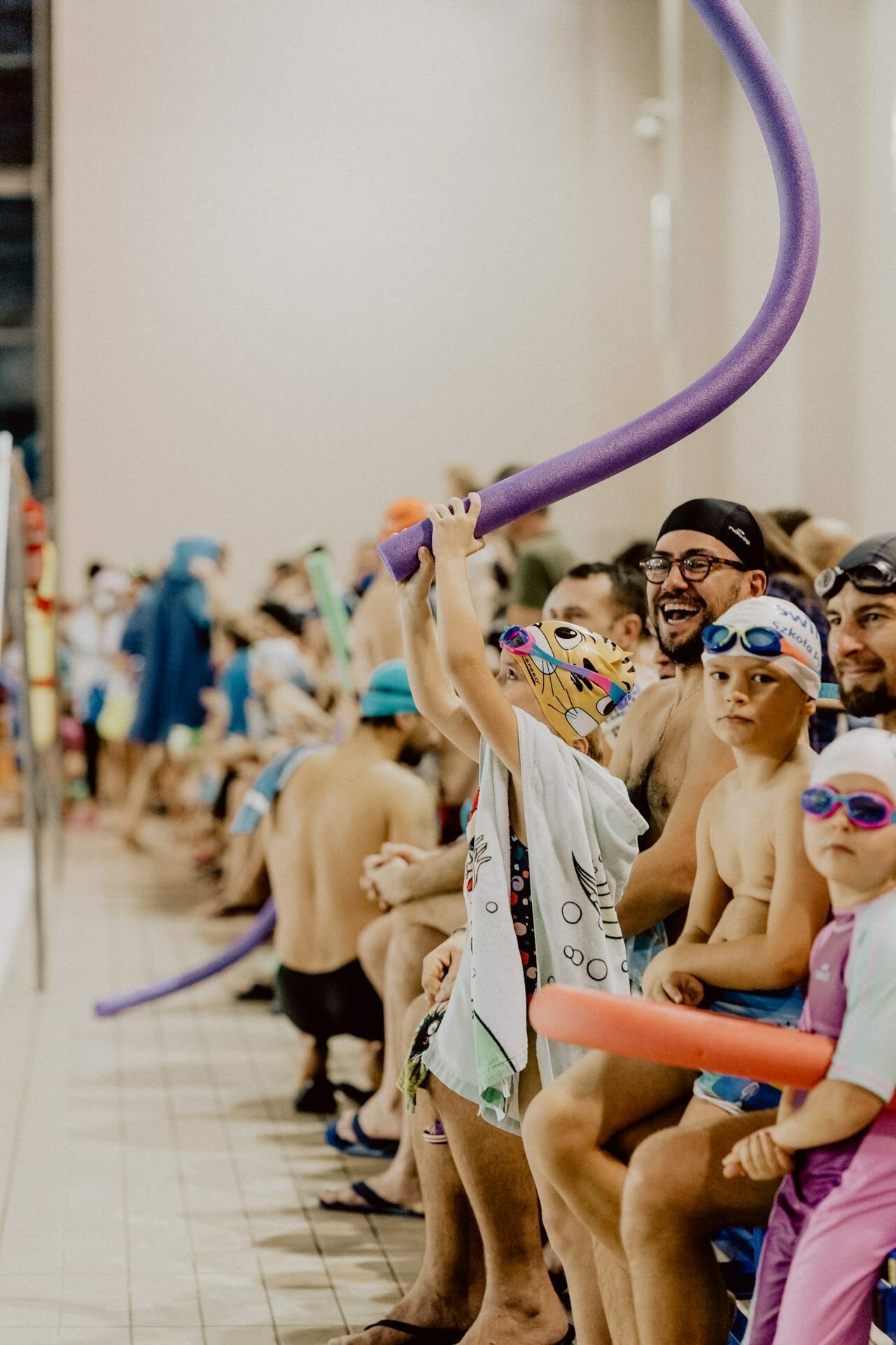 A group of people, including children and adults, are sitting on the edge of an indoor pool. They are wearing bathing suits and swimming goggles. A child in the foreground playfully holds a purple pool noodle. In the background, more swimmers and parents can be seen sitting and waiting. This photo captures the essence of event photography, as exemplified by the skillful event photojournalism by Marcin Krokowski.    