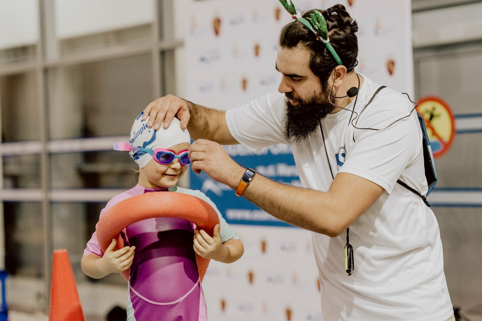 A bearded man wearing a headband adjusts the swimming goggles of a young child wearing a pink bathing suit and swim cap. The child holds a red pool noodle and stands near the pool, ready for a swimming lesson. In the background are banners and logos, capturing the essence of event photography.  