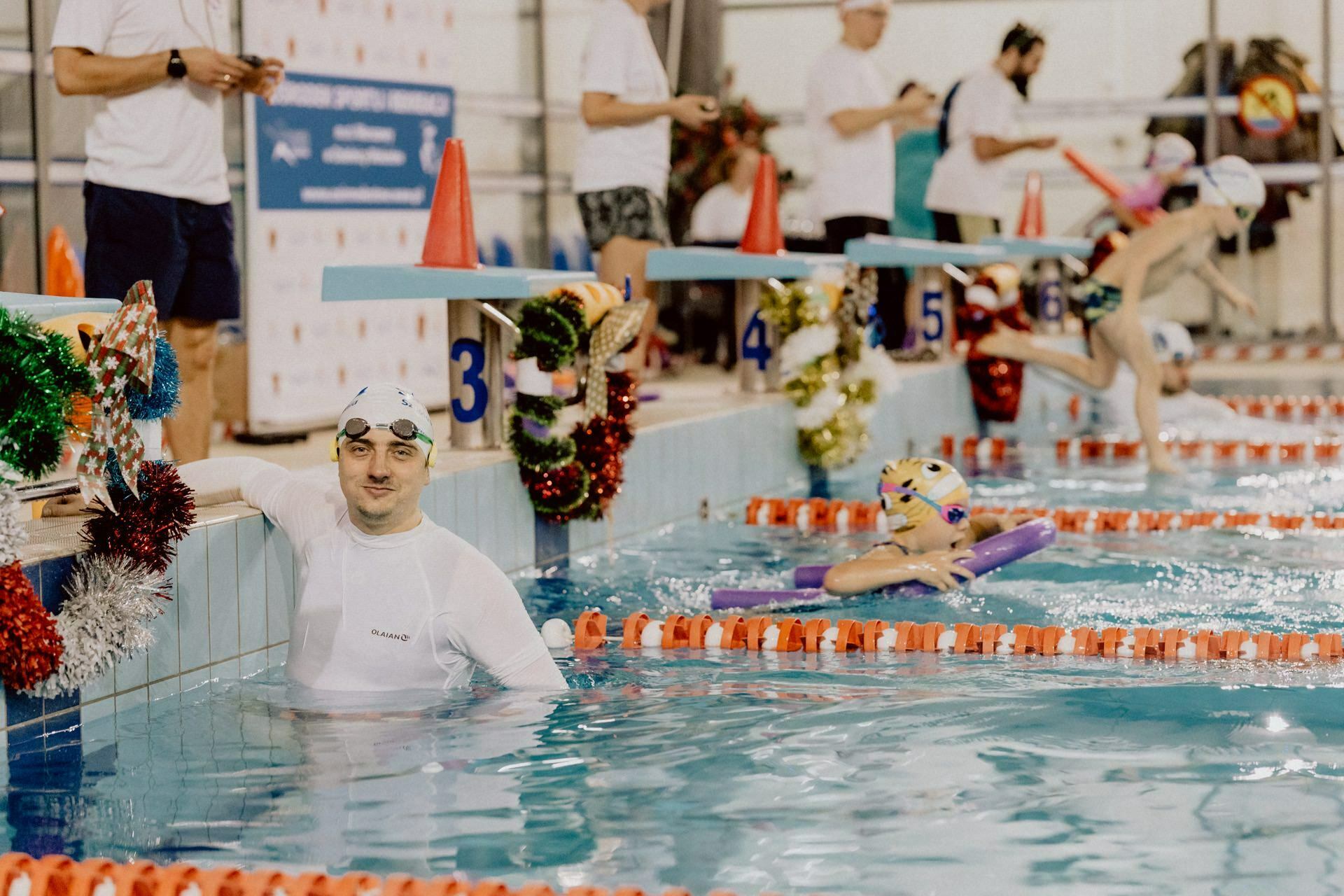 Swimmers prepare for a race at the starting blocks of an indoor pool. One swimmer in a white cap and costume waits in the water, while another in a colorful cap warms up next to them. The pool area was decorated with festive garlands, which were perfectly captured by Marcin Krokowski's photo report of the events.  