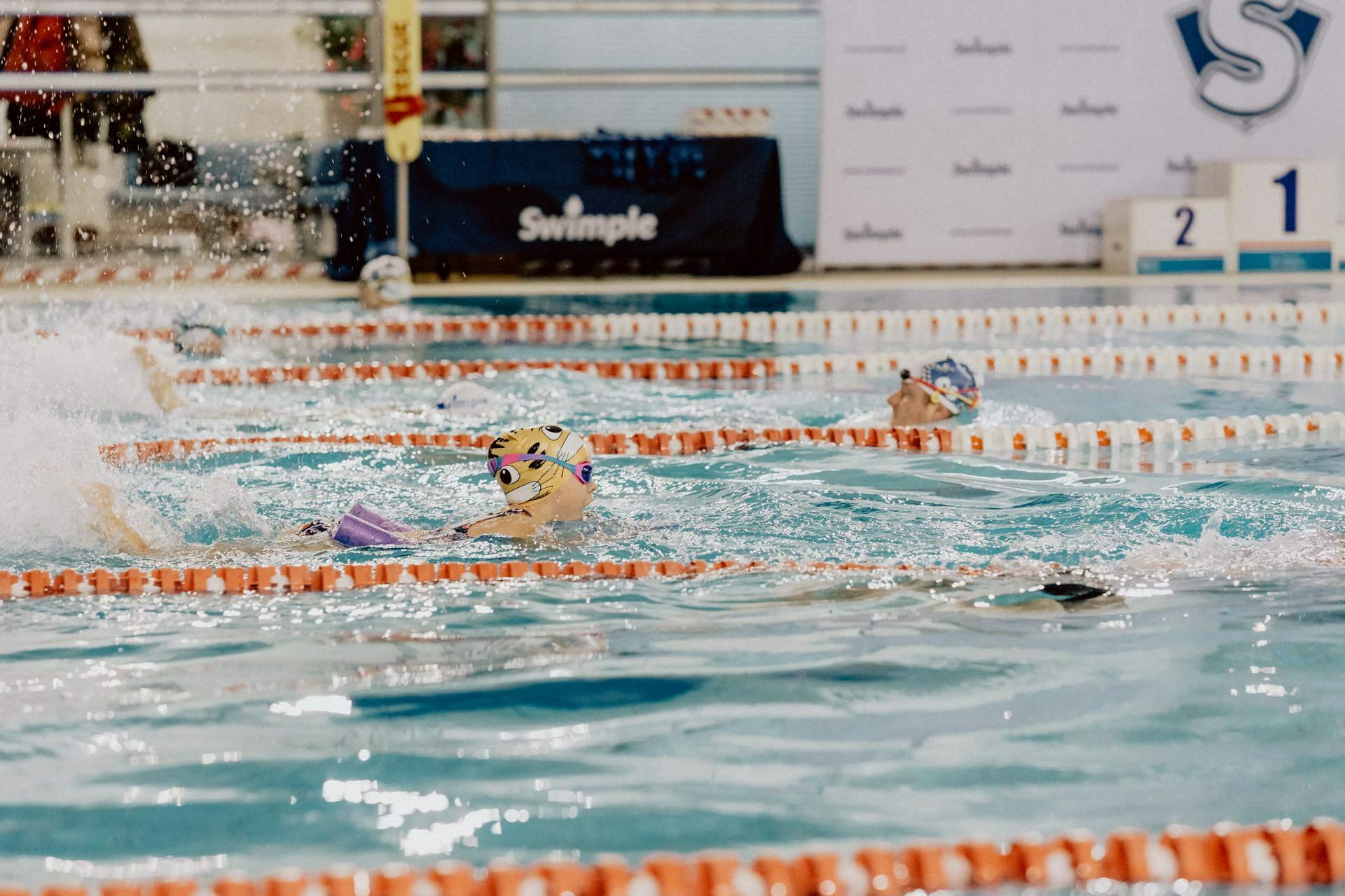 A group of swimmers compete in an indoor pool race, near the edge of the course divider, with water splashing vigorously around them. In the background, a banner with logos and a podium with numbered stands, perfectly captured by Marcin Krokowski's event photography. 