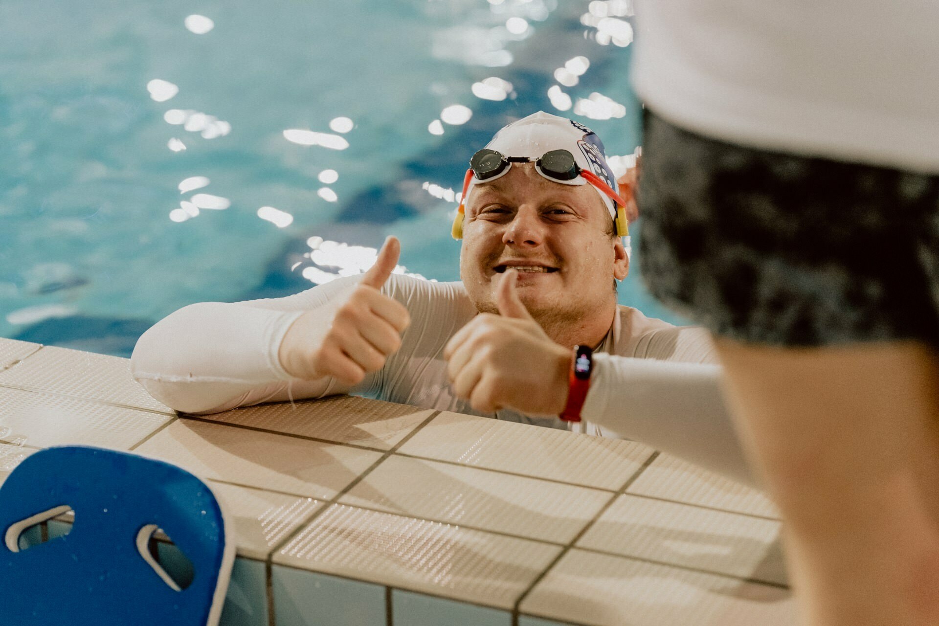 A swimmer wearing a cap and goggles is in the pool, resting his hands on the edge. He shows two thumbs up and smiles at the camera. Another person's lower body is visible nearby. The water in the pool is a vivid blue, perfectly captured by Marcin Krokowski, known for his expert event photography.   