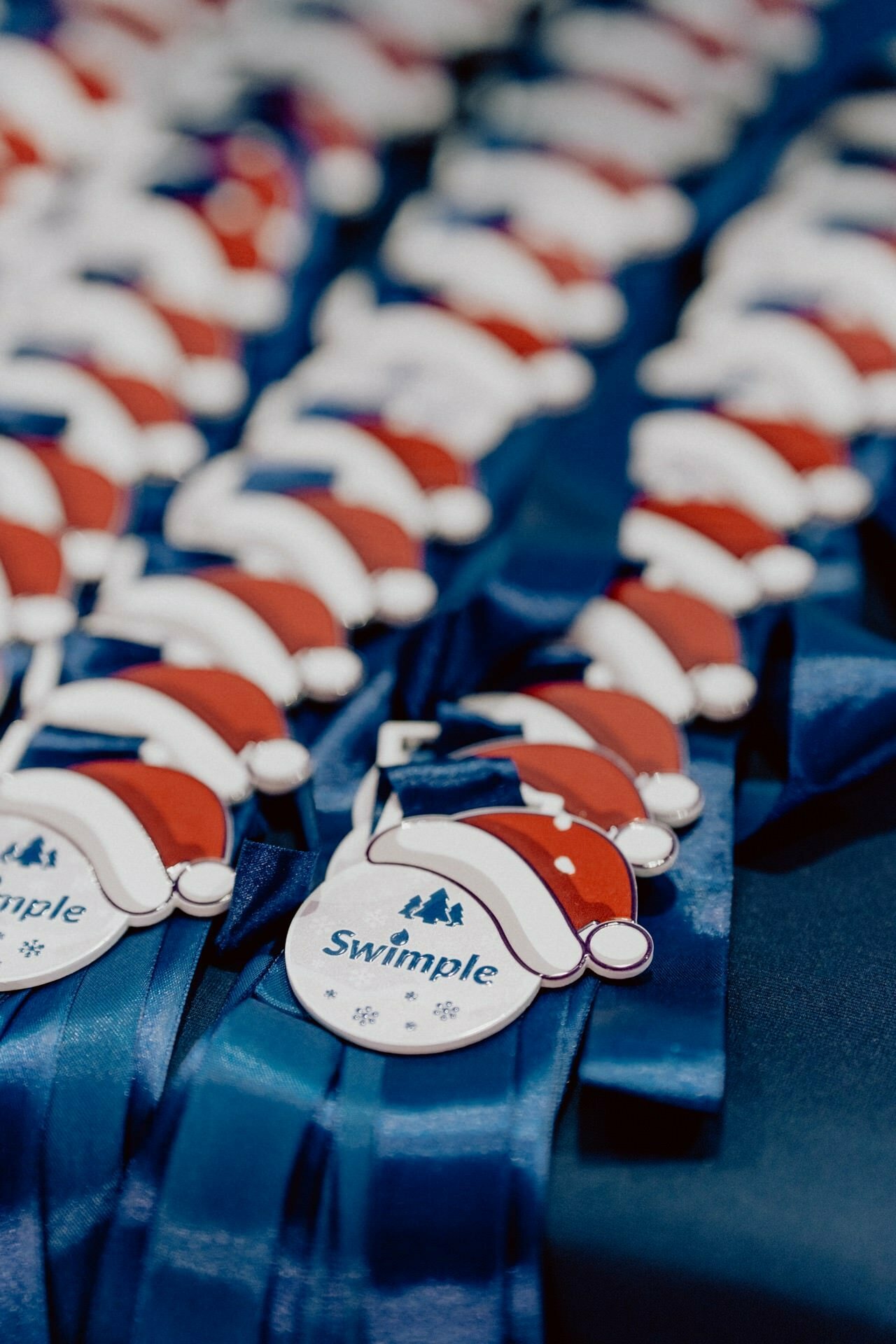 Rows of medals with blue ribbons and a red Santa hat design, decorated with the word "Swimple," are neatly arranged on a table. The photo, taken by Marcin Krokowski, could be part of a Christmas or holiday swimming event photo essay. 