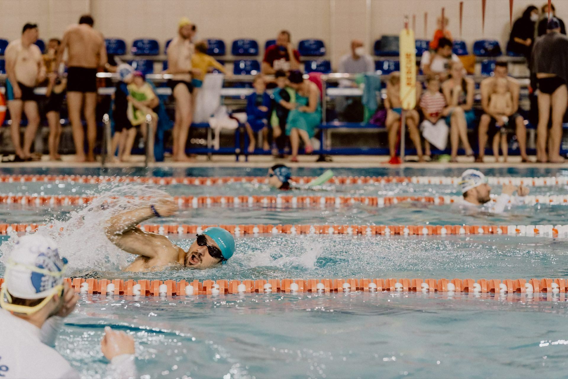 A swimmer wearing goggles and a swimming cap competes in a freestyle race in an indoor pool. Other swimmers and spectators can be seen in the background, some sitting on benches, others standing. The event photo captures the pool divided by track markers, highlighting the competitive atmosphere.  