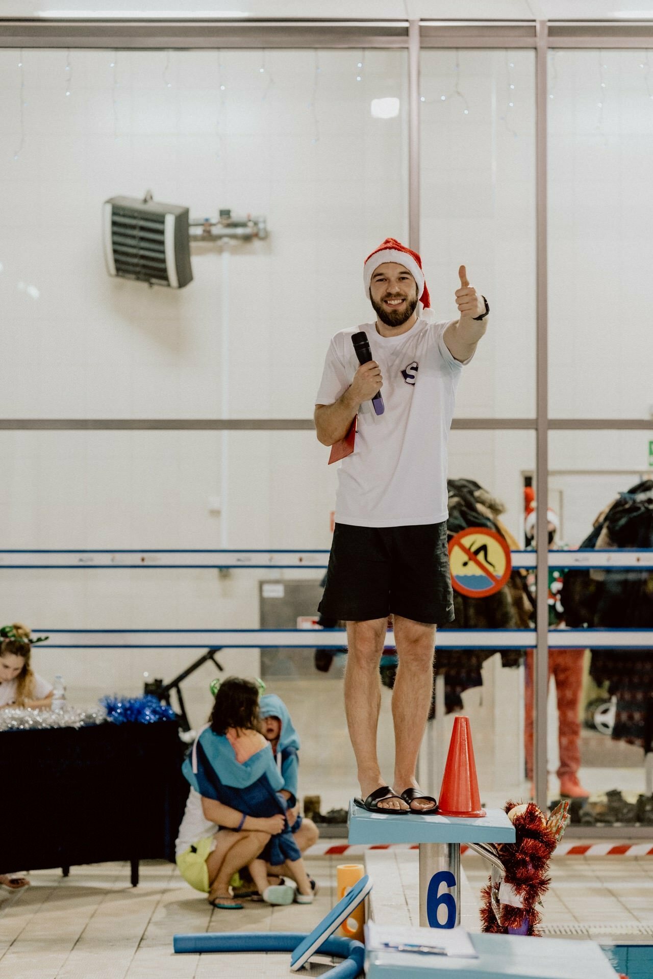 A man in a Santa hat and white shirt stands on a platform next to the pool and shows thumbs up. In his other hand he holds a microphone and smiles. In the background are swimmers, some seated, against a backdrop of windows and swimming equipment. The scene was captured by Marcin Krokowski, a Warsaw-based event photographer.   