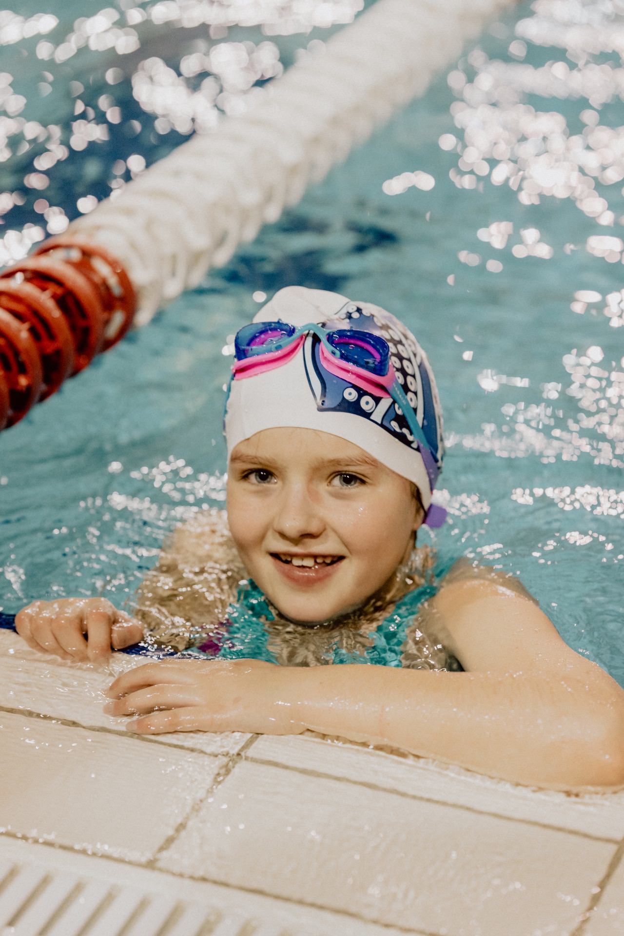 A smiling child wearing colorful swimming goggles and a swimming cap rests on the edge of the pool, holding onto the side with one hand. The water is clear, and the other lane is parallel to them, separated by a red and white divider, which was beautifully captured by event photographer Marcin Krokowski. 