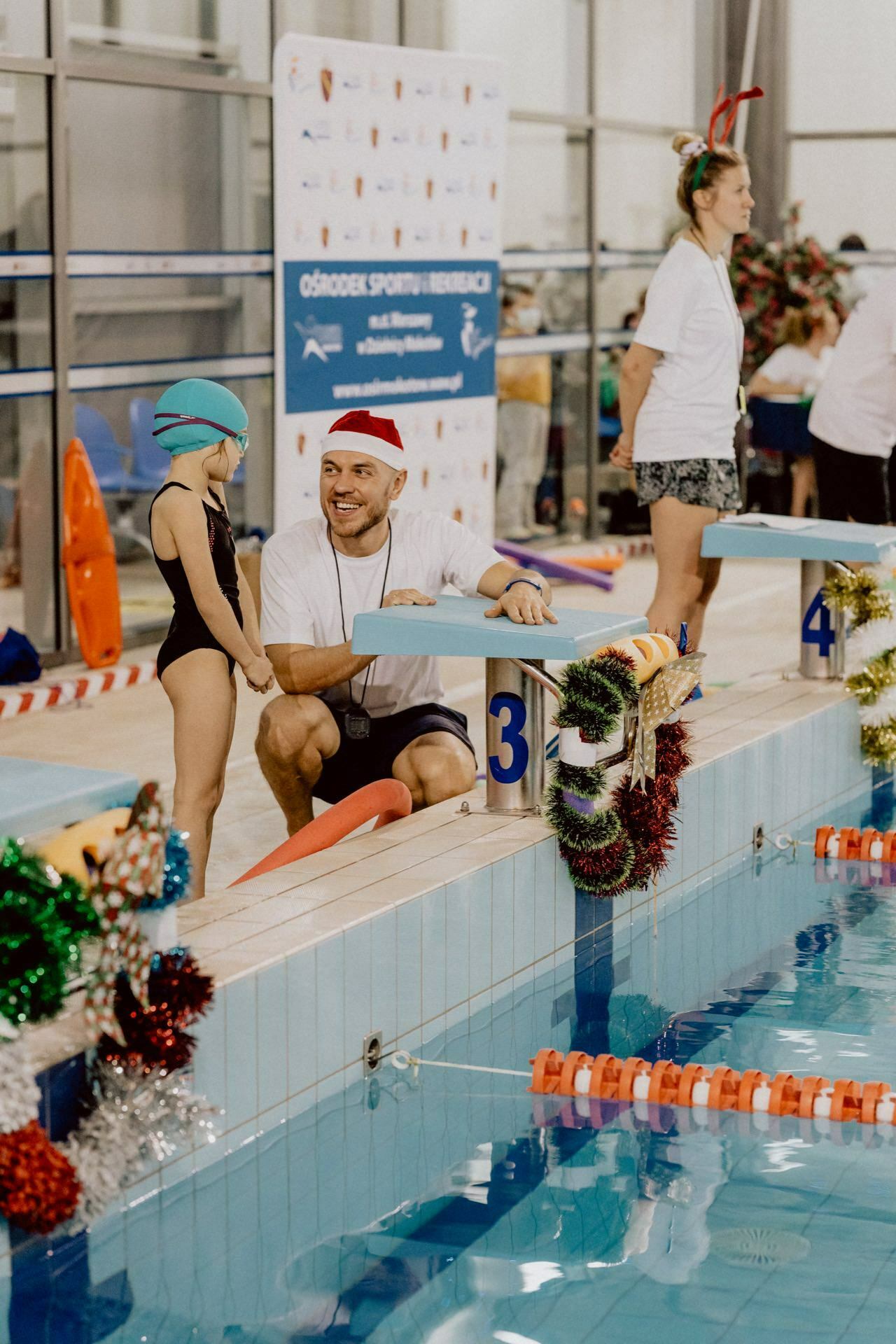 A child in a cap and bathing suit stands by the pool and talks to an adult in a Santa hat and white shirt, kneeling by the pool. The pool area is decorated with Christmas lights. A banner and other people are visible in the background, capturing the delightful event photography.  