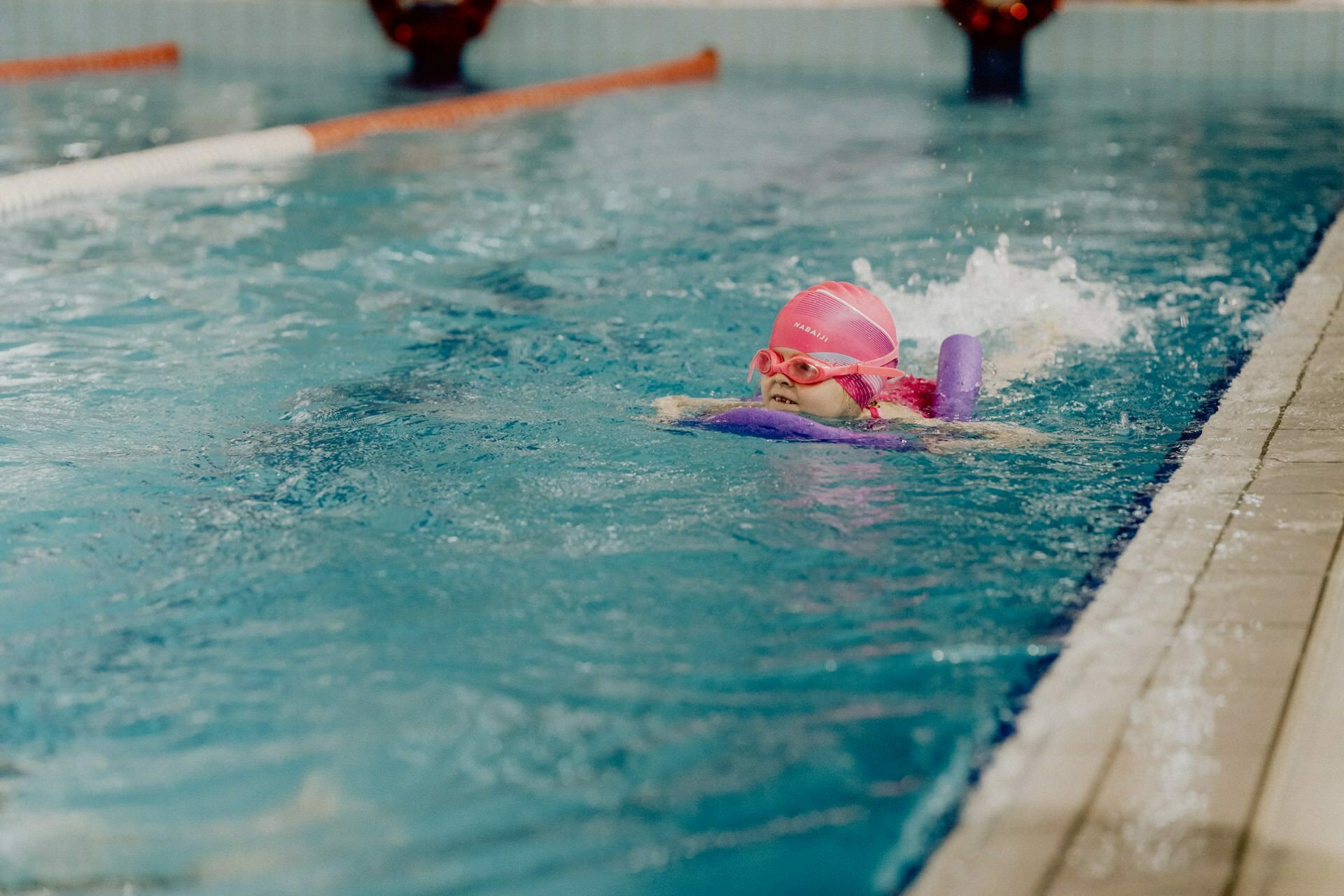 A young child wearing a pink cap and goggles is practicing swimming in a pool. Guided by a board, they move near the edge of the pool, creating splashes of water around them. This moment was beautifully captured by Marcin Krokowski, a talented event photographer from Warsaw.  