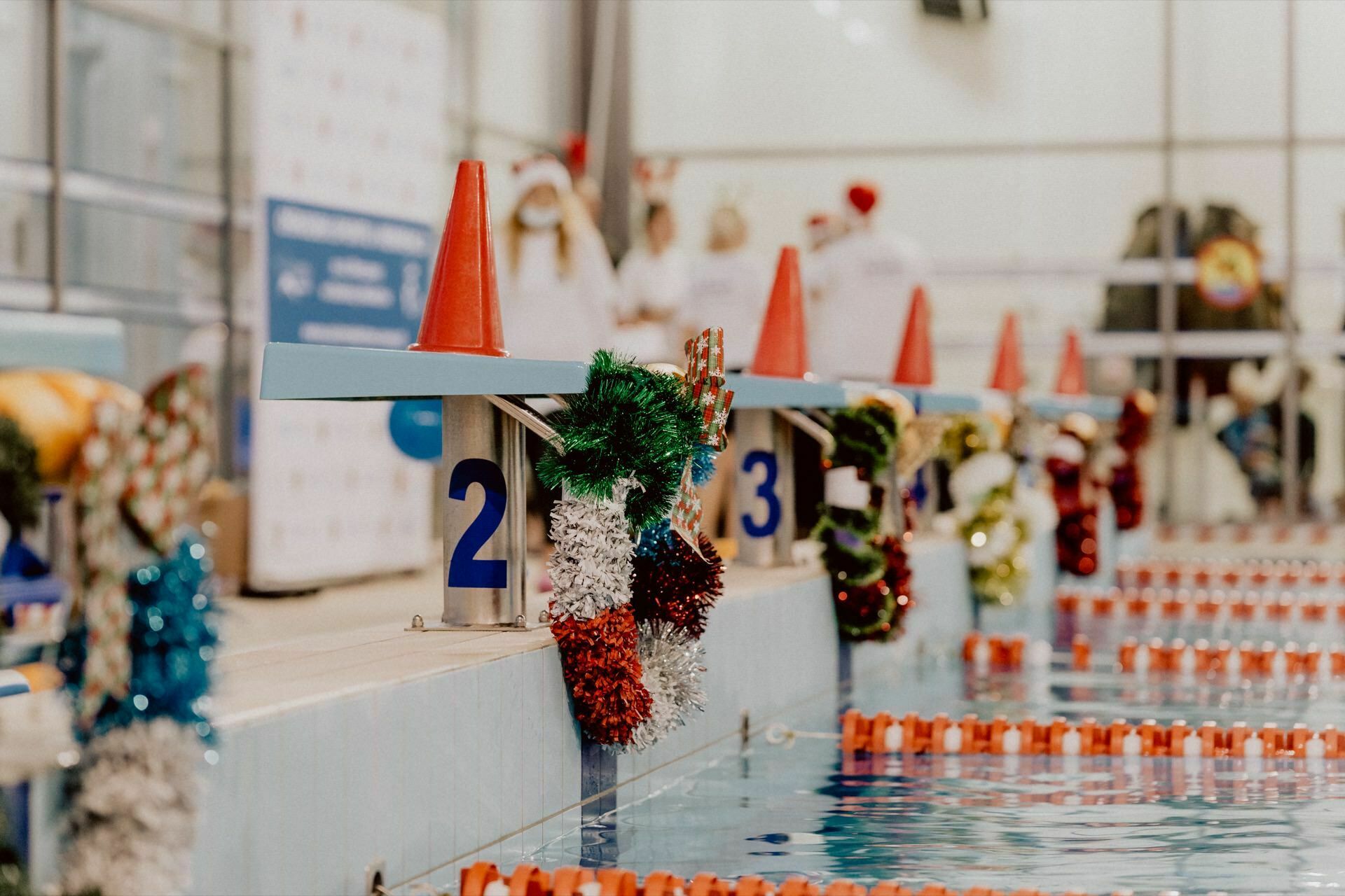 The starting blocks at the pool are decorated with Christmas ornaments and candles. The tracks are marked with orange track dividers, and the numbers on the blocks are clearly visible. The people in the background are out of focus, capturing the lively atmosphere of the photo event.  