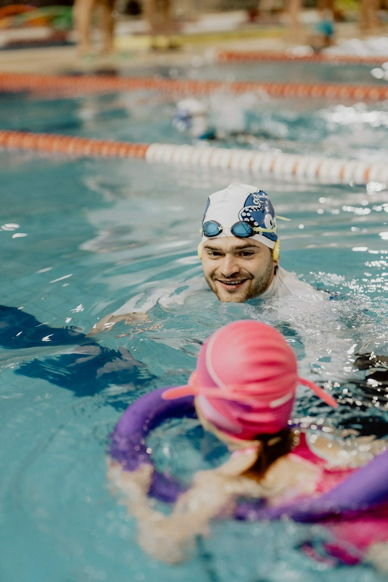 A swimming instructor wearing a cap and goggles smiles as he interacts with a young child wearing a pink cap and swimming pool equipment. The child uses a purple floatation device. Other swimmers and colorful lane splitters can be seen in the background - a delightful photo report of the events captured by Marcin Krokowski.  