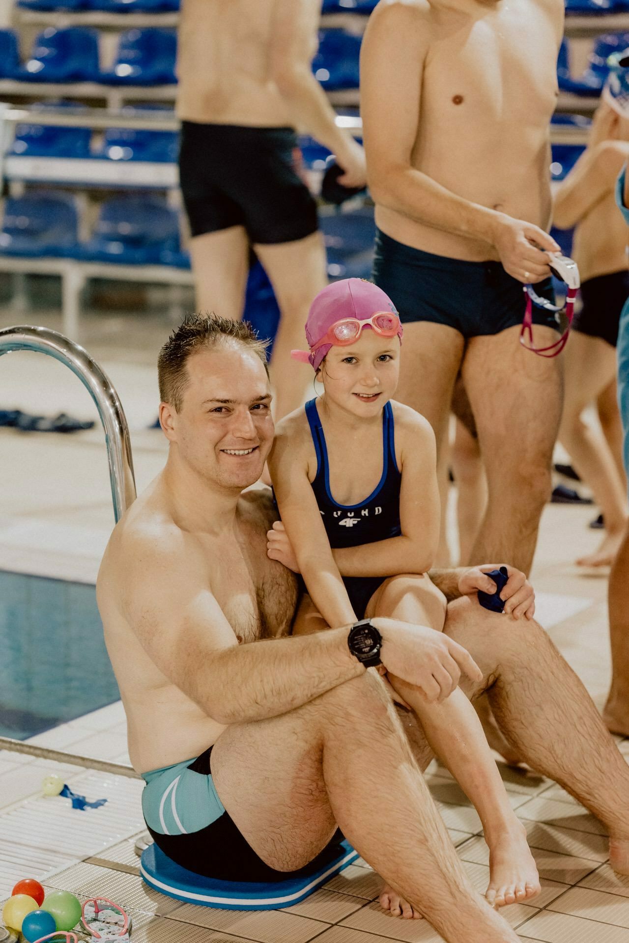 A man in swimwear sits by the pool with a young girl on his lap, who is wearing a pink swim cap and goggles. In the background near the pool are other people in bathing suits. Toys are scattered on the deck. The two are smiling and look happy, capturing an uplifting moment, perfect for an event photo essay.   