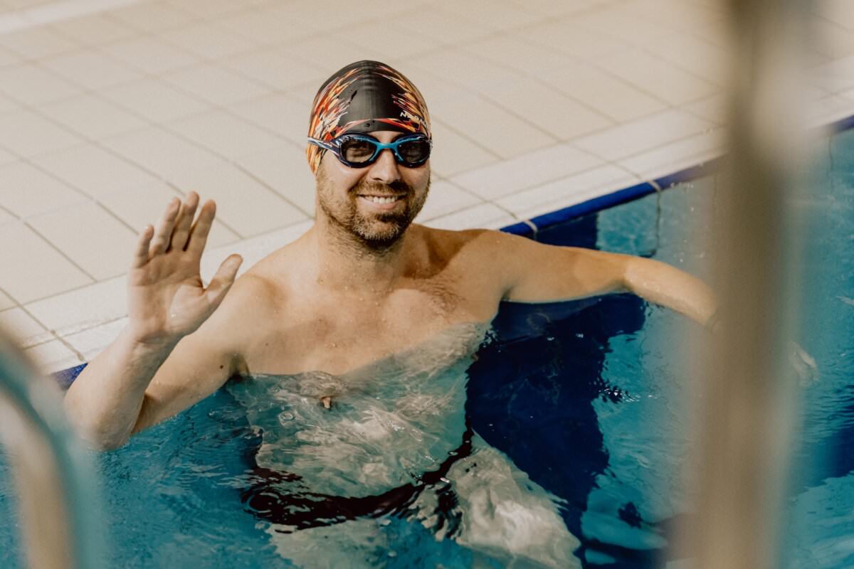 Uśmiechnięty mężczyzna w czepku i okularach pływackich macha do basenu. Odpoczywa na brzegu basenu, częściowo zanurzony w wodzie. W tle widoczne płytki basenowe – idealne ujęcie do fotorelacji z wydarzenia.