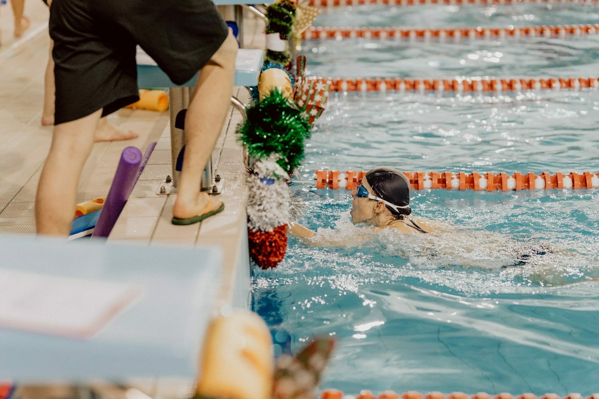 A swimmer wearing a swimming cap and goggles touches the wall of the pool lane during a swimming competition. An official standing on a nearby terrace by the pool appears to be watching the swimmer. The pool, captured by photographer Marcin Krokowski, is decorated with festive garlands and orange partitions.  