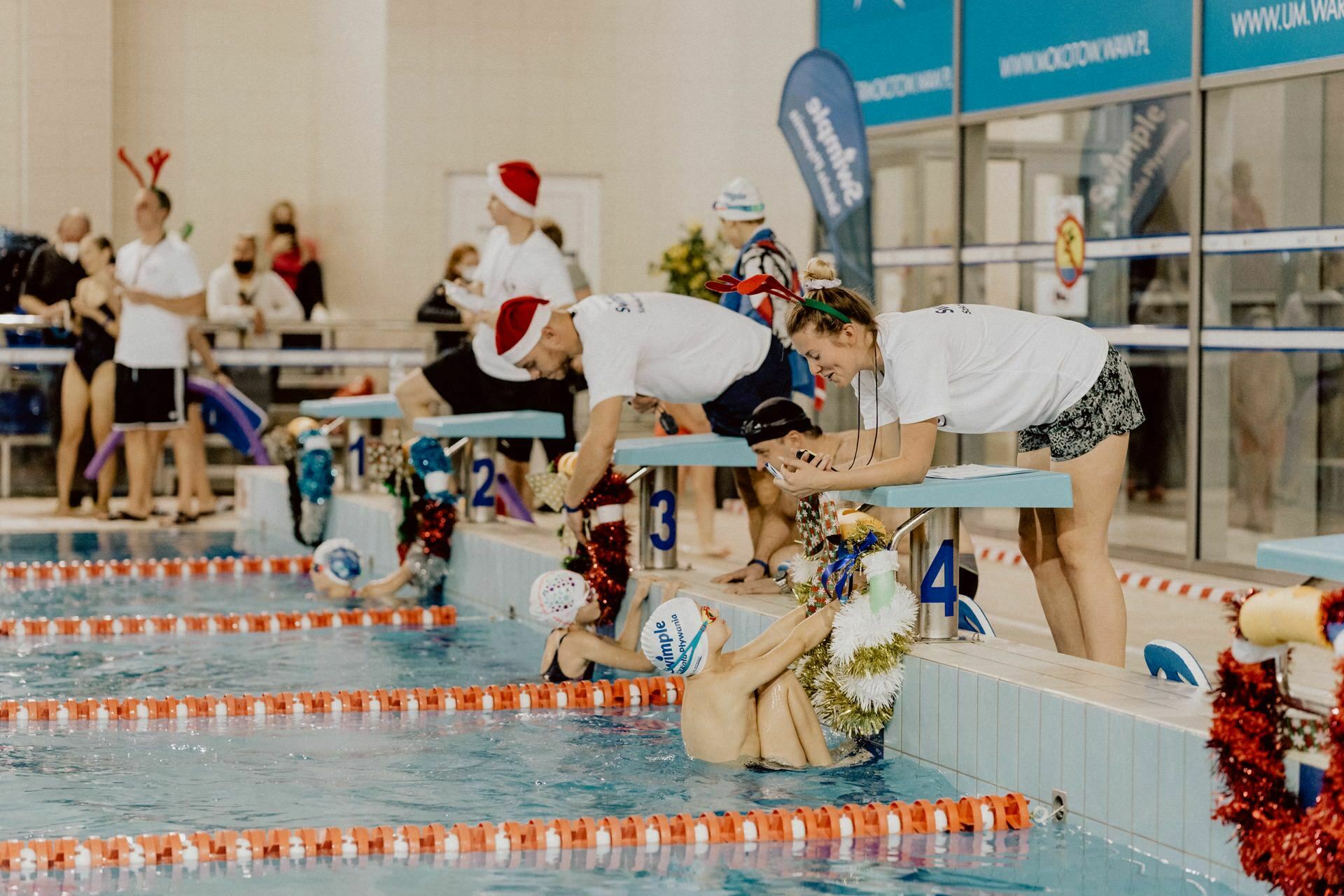 Swimmers in the pool prepare for a race wearing swimming caps and goggles, while coaches lean over the starting blocks giving instructions. Christmas decorations and people in festive paraphernalia suggest a holiday-themed event, perfect for event photography by event photographer warsaw. 