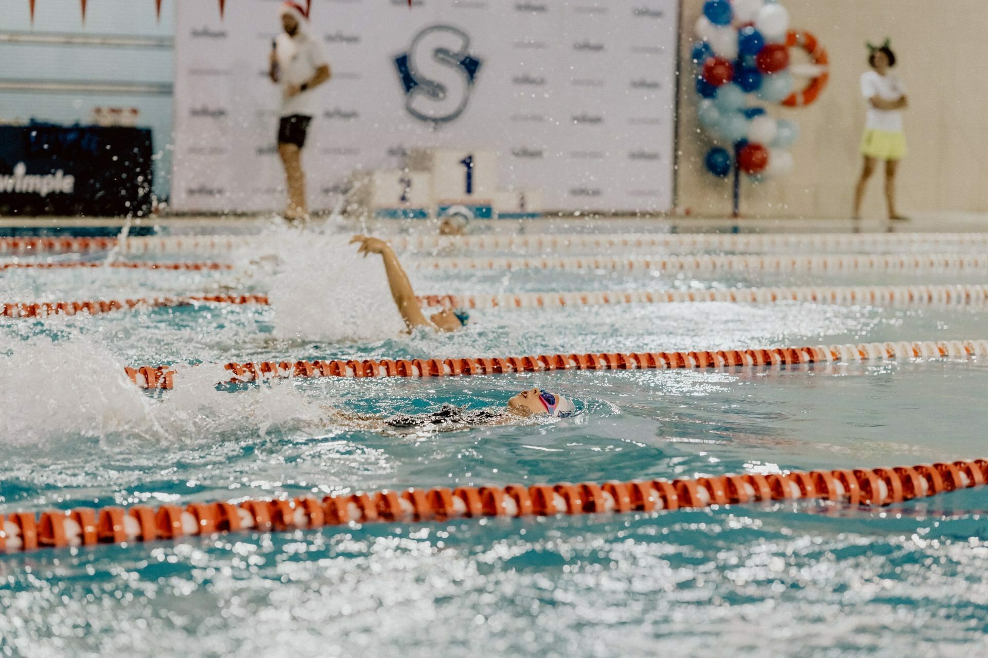 Swimmers taking part in the race form spots as they move along the lanes of the indoor pool. In the background, an audience is visible against a decorated wall with a large "S" logo and colorful balloons. The atmosphere, captured by event photography, is lively and focused on the event.  