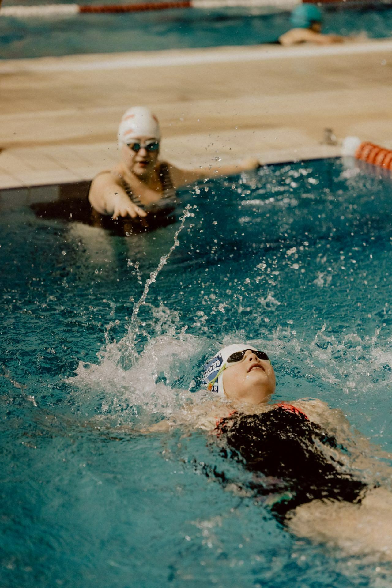 A swimmer in a black swimsuit with red straps and a white swimming cap performs a backstroke, creating a splash around him. Another swimmer in a black swimsuit and white swim cap stands at the edge of the pool and watches. This photo recap of the events shows the intensity and focus of the swimming competition.  