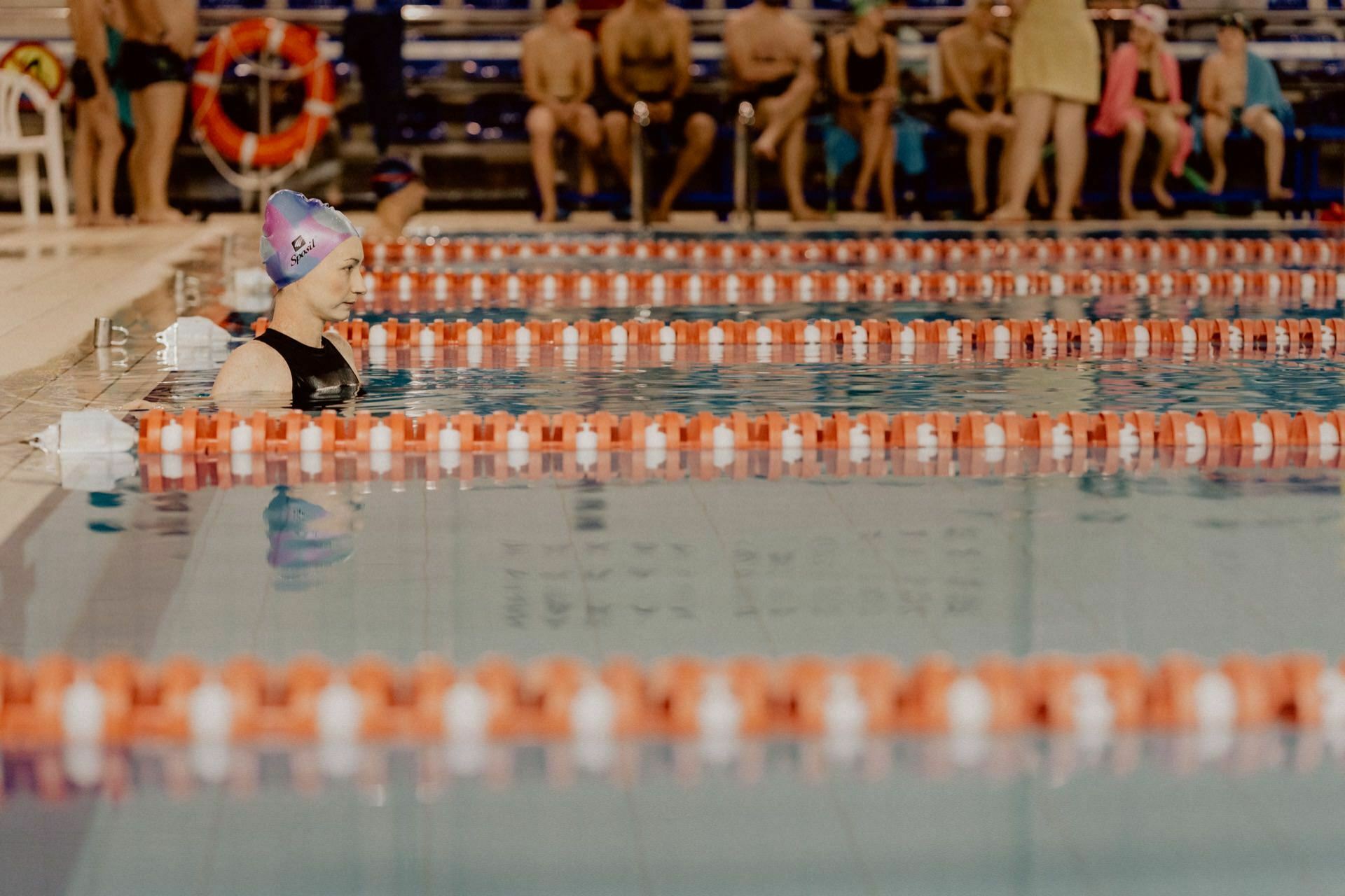 A swimmer wearing a purple cap and black bathing suit stands on a track at the edge of an indoor pool. The pool is divided by orange and white dividers. Several people sit on benches in the background and watch, making for fascinating event photography.  