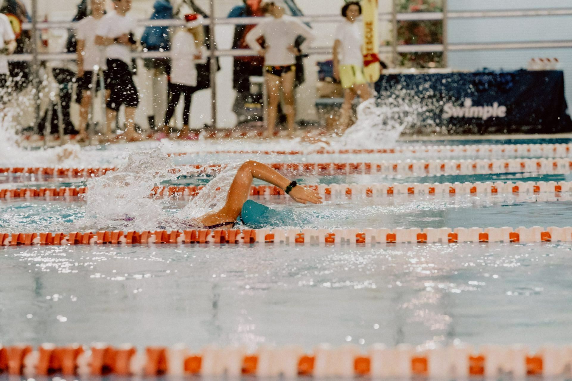 A swimmer takes part in a freestyle race, splashing water and swimming forward on a course marked with orange and white swimmers. In the background, spectators and swimmers in white outfits stand by the pool, as brilliantly captured by Marcin Krokowski in a photo report of the events. 