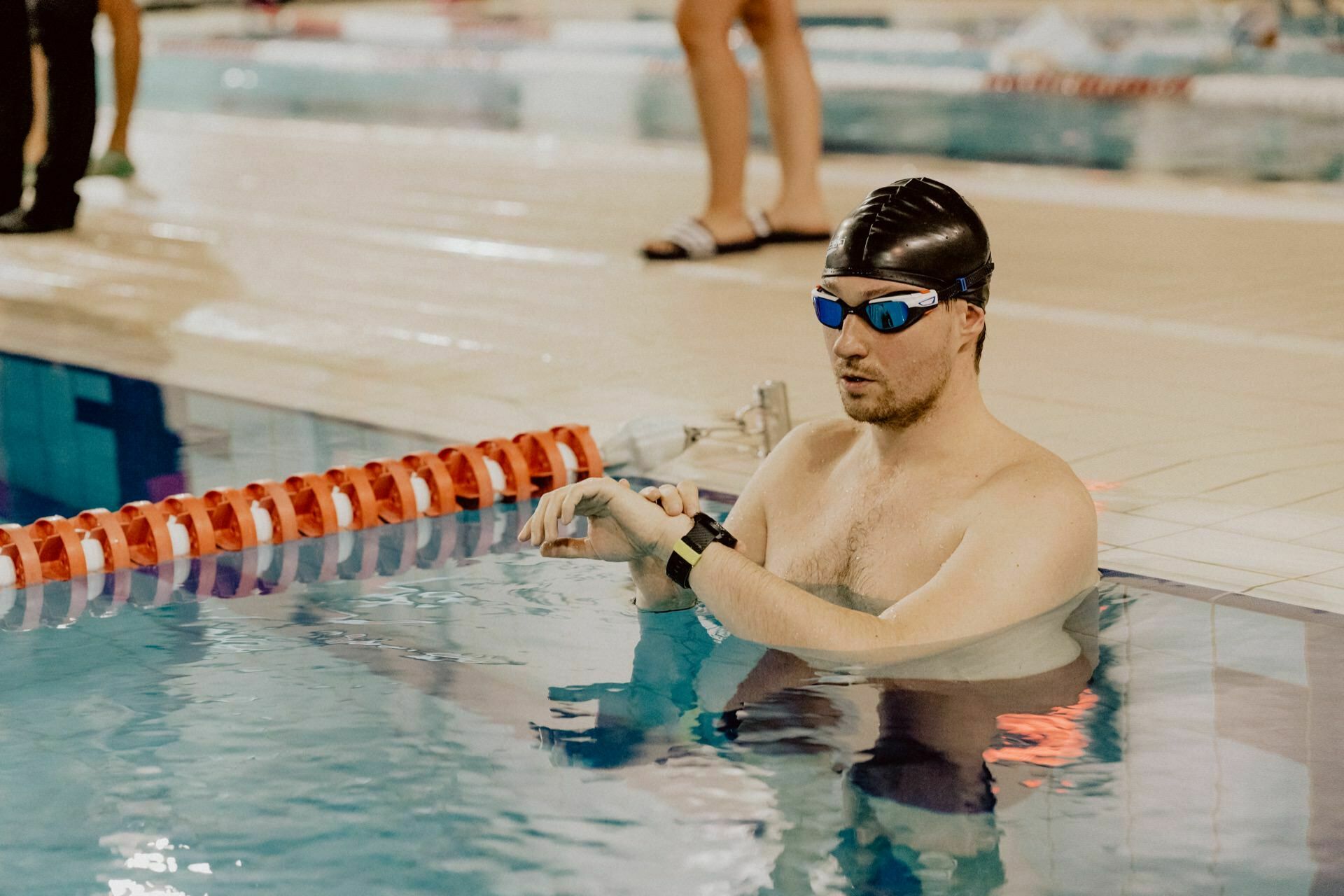 A swimmer wearing a black cap and blue goggles rests on the edge of the pool and looks at his watch. Red and white belt splitters are visible in the water, and a blurry person stands in the background on the terrace by the pool. Capture this moment perfectly with the expertise of Marcin Krokowski.  