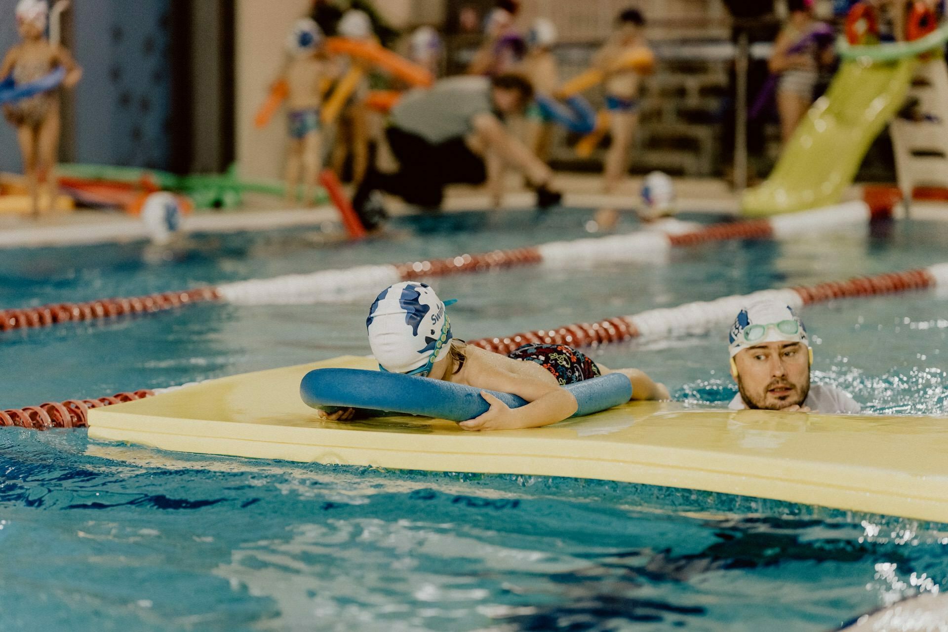 A child wearing a swimming cap and goggles holds onto a foam board while swimming in the pool, accompanied by an adult swimmer who guides them. Other children and adults are in the background, close to the edge of the pool, with various pool equipment visible, capturing the lively event photography. 