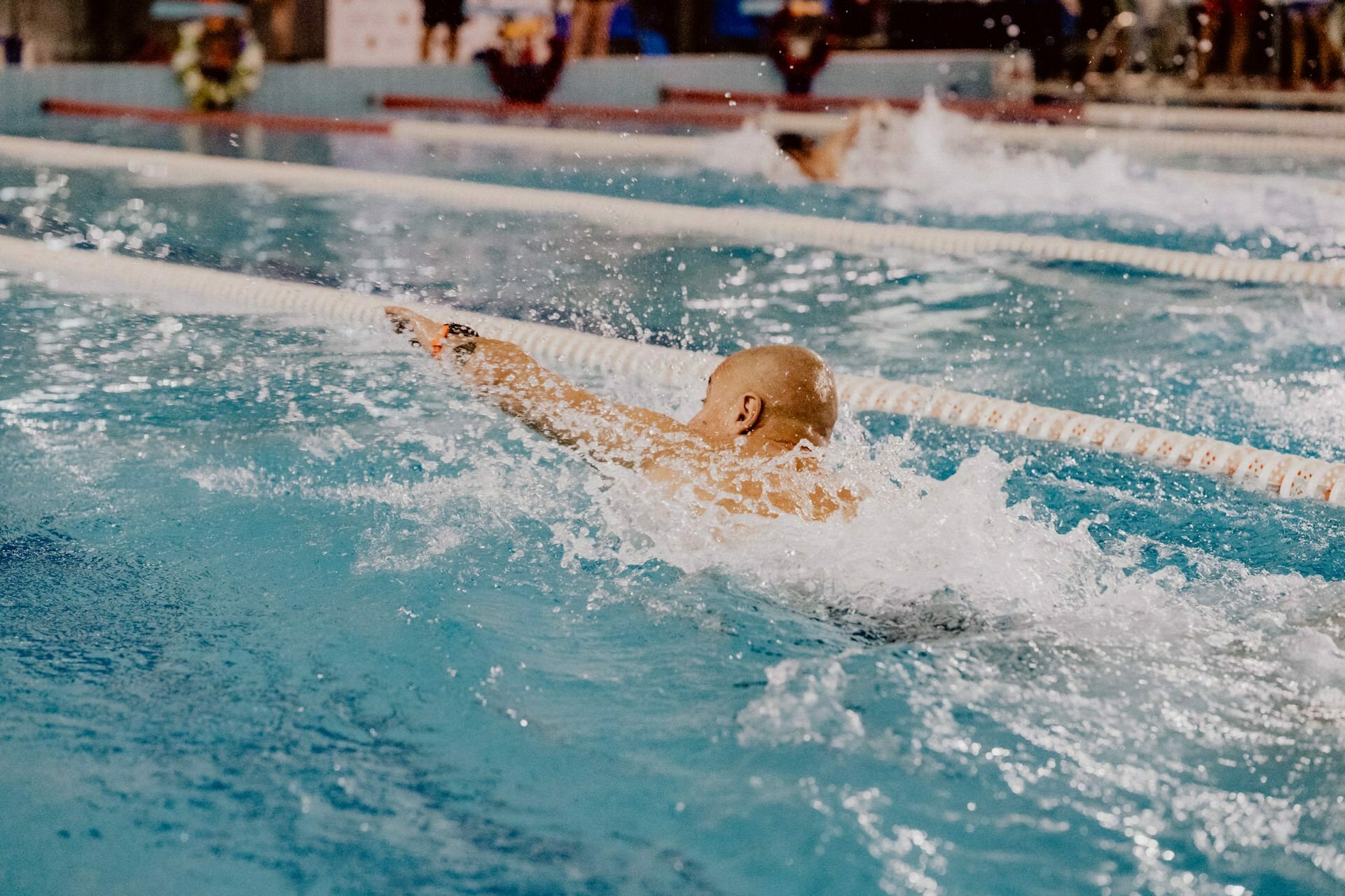 A bald-headed swimmer is seen in the pool in mid-motion, with white baffles on either side. Water splashes around them as they walk forward. Other swimmers can also be seen in the adjacent lanes, as perfectly captured by the photo report of the events.  