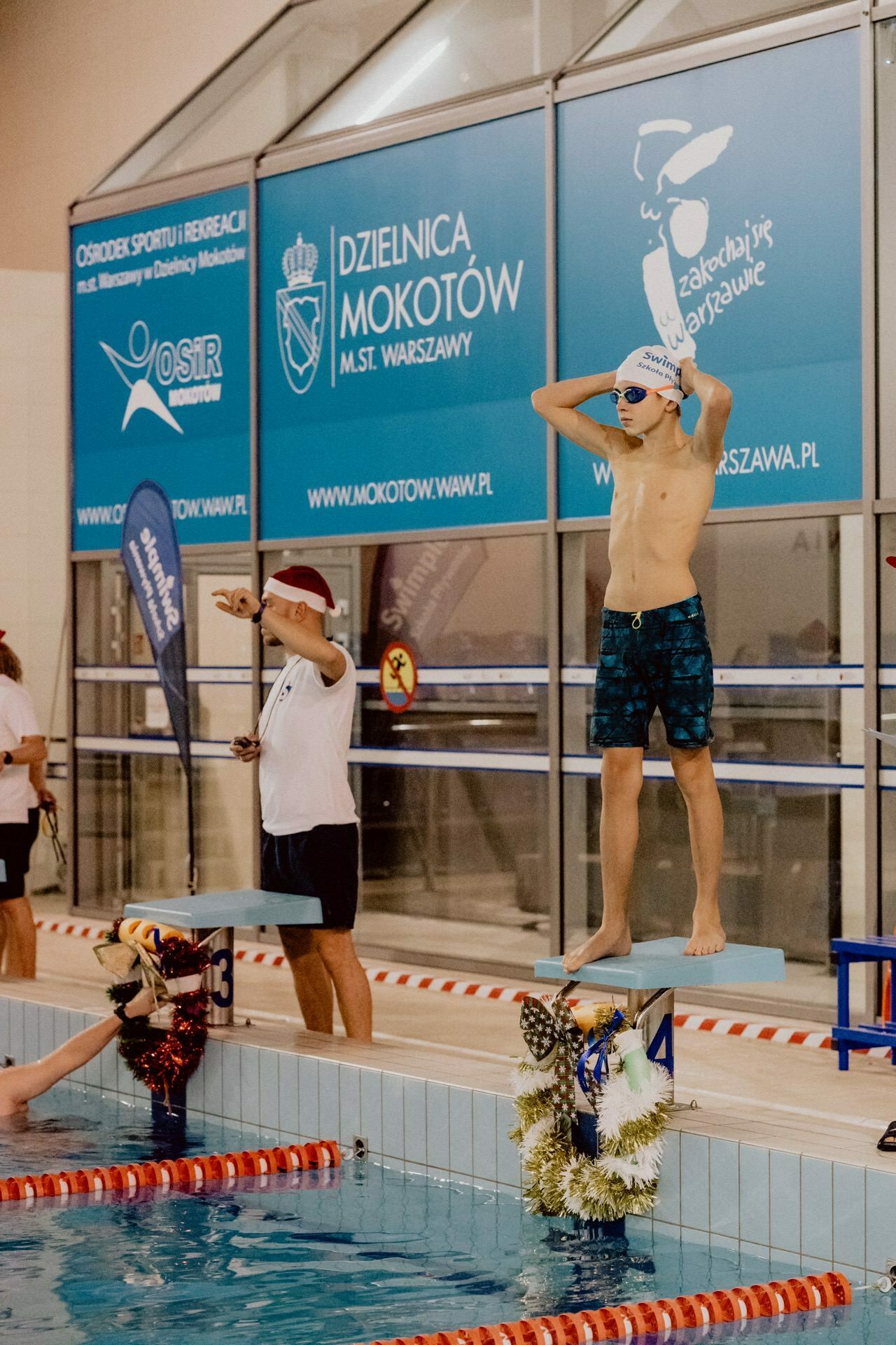 A swimmer in swim trunks and cap stands on the diving block, tweaks his goggles and prepares to dive into the pool. Next to him, an official wearing a Santa hat signals with a whistle. The signage of the event behind them indicates that it takes place in Warsaw's Mokotow district. Photo coverage of the event vividly captures every moment.   