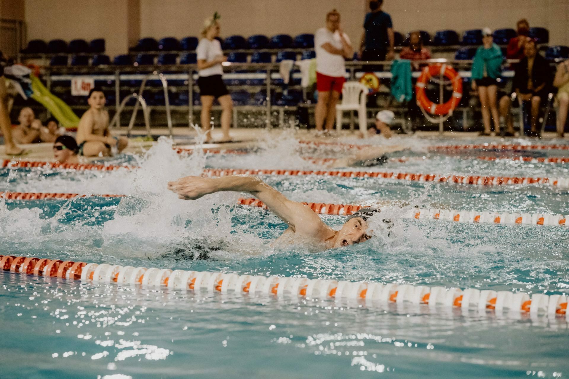 A swimmer takes part in a freestyle race in an indoor pool, splashing water as he moves. Spectators and other swimmers can be seen in the background. The pool is lined with orange and white markers that capture the action for event photography enthusiasts.  