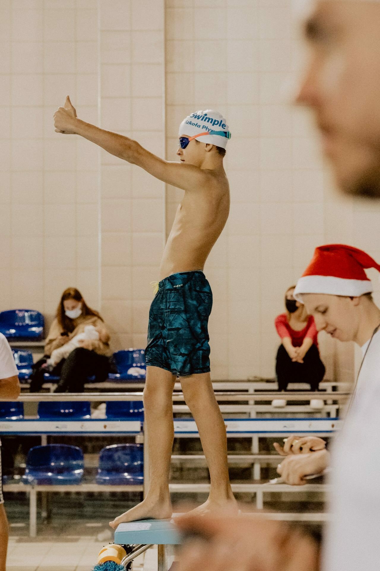 A young swimmer stands on a diving block, one hand showing a thumb up. He wears a swimming cap, goggles and swim trunks. In the background, people are sitting in the stands, some wearing masks. The person in the foreground is partially visible, wearing a Santa hat. This vibrant scene is part of a photo essay of the event showing the excitement of the competition.    