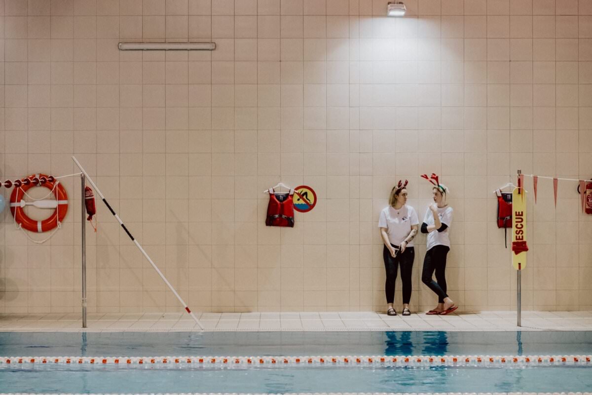 Dwie osoby ubrane w zwykłe stroje sportowe stoją przy basenie w krytym obiekcie pływackim. Prowadzą rozmowę, a na ścianie za nimi widoczne są kamizelki ratowników, koła ratunkowe i słupek zabezpieczający. Basen wyposażony jest w pływające przegrody, które idealnie nadają się do uchwycenia momentów związanych z wydarzeniami fotograficznymi.