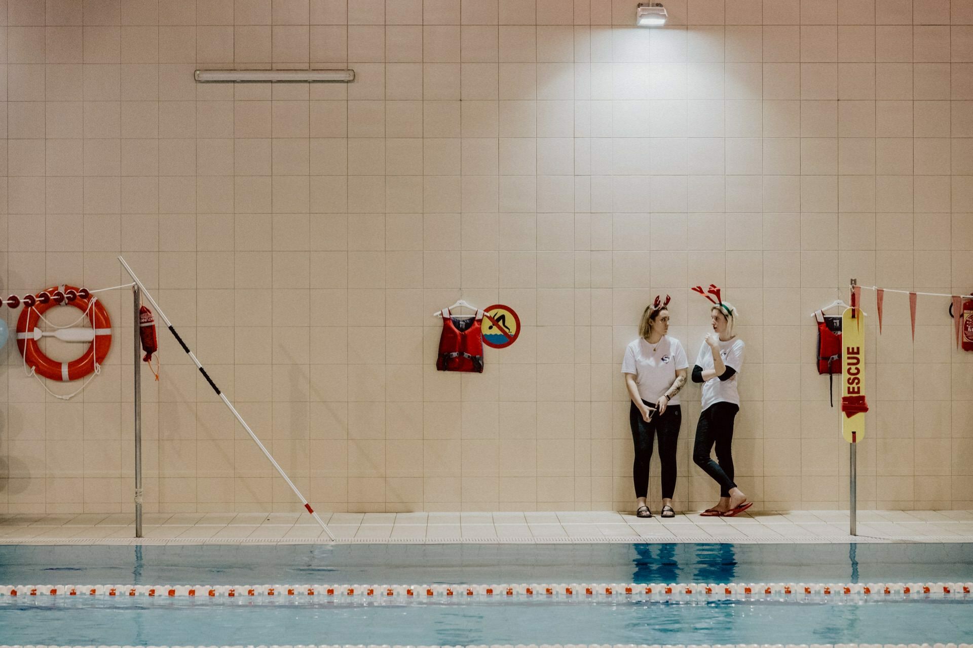 Two people dressed in casual sportswear are standing by the pool at an indoor swimming facility. They are engaged in a conversation, while lifeguard vests, lifebuoys and a safety post are visible on the wall behind them. The pool is equipped with floating partitions, which are ideal for capturing moments of photographic events.  