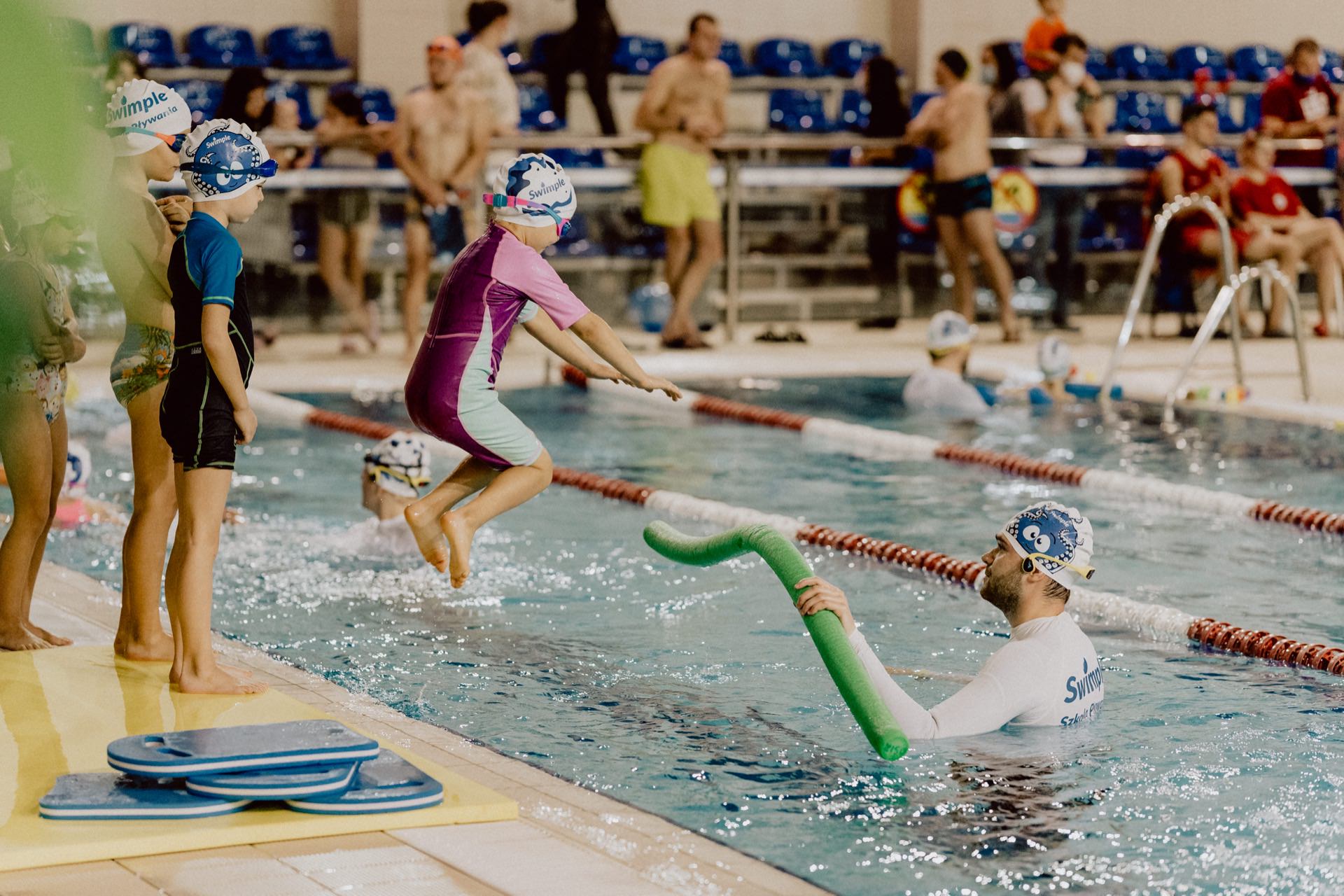 Children wearing swimming caps and goggles take part in a swimming class at an indoor pool. One child in a pink swimsuit jumps into the water toward an instructor holding a green pool noodle. More children and adults can be seen in the background, near the grandstands and pool terrace. This scene was captured by event photographer Marcin Krokowski for the event photo report.   