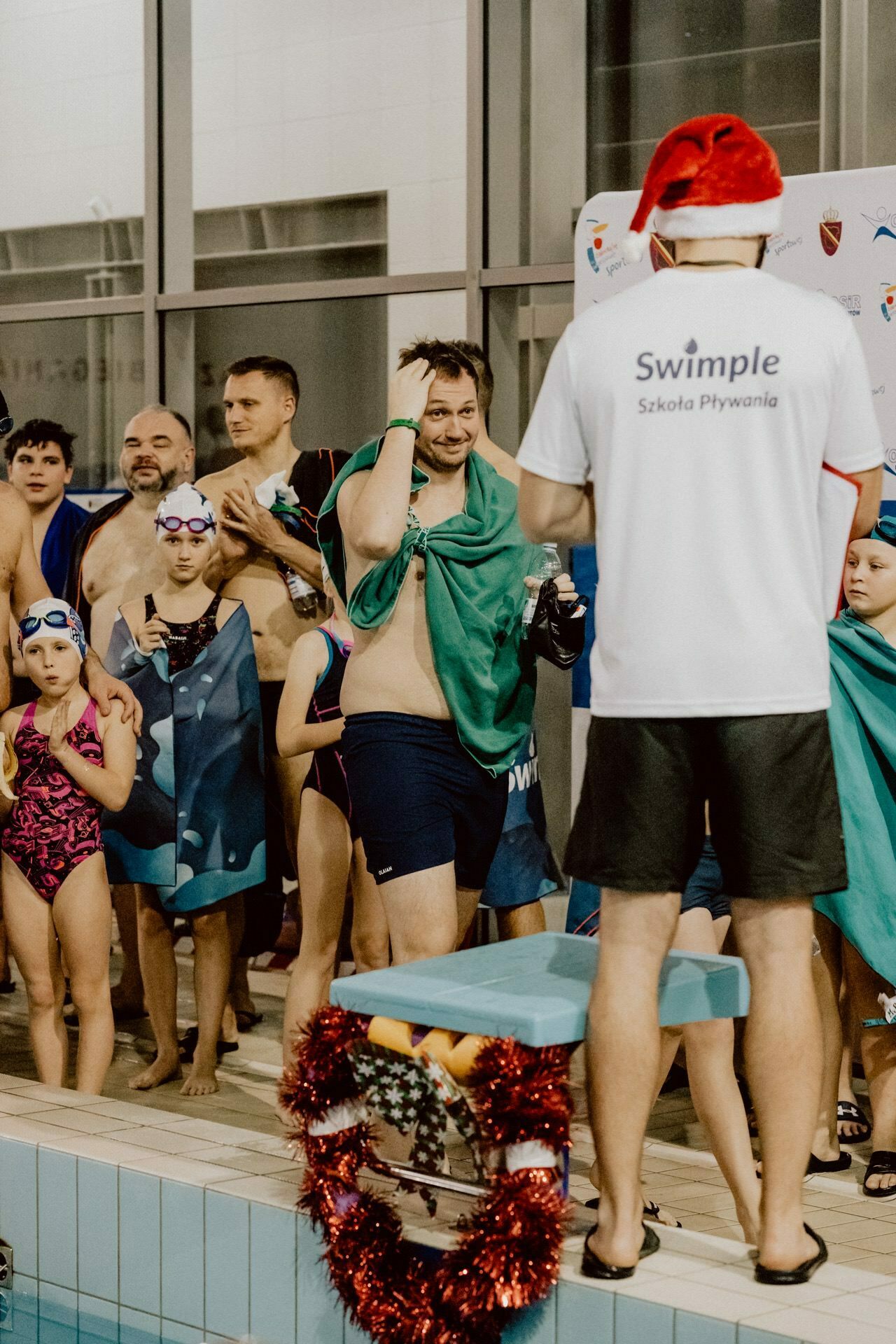 A group of swimmers, both children and adults, stand by the pool in bathing suits and towels and listen to a person wearing a Santa hat and a white "Swimple Swim School" T-shirt. A Christmas wreath adorns the platform in the foreground, capturing the spirit of this photo event. 