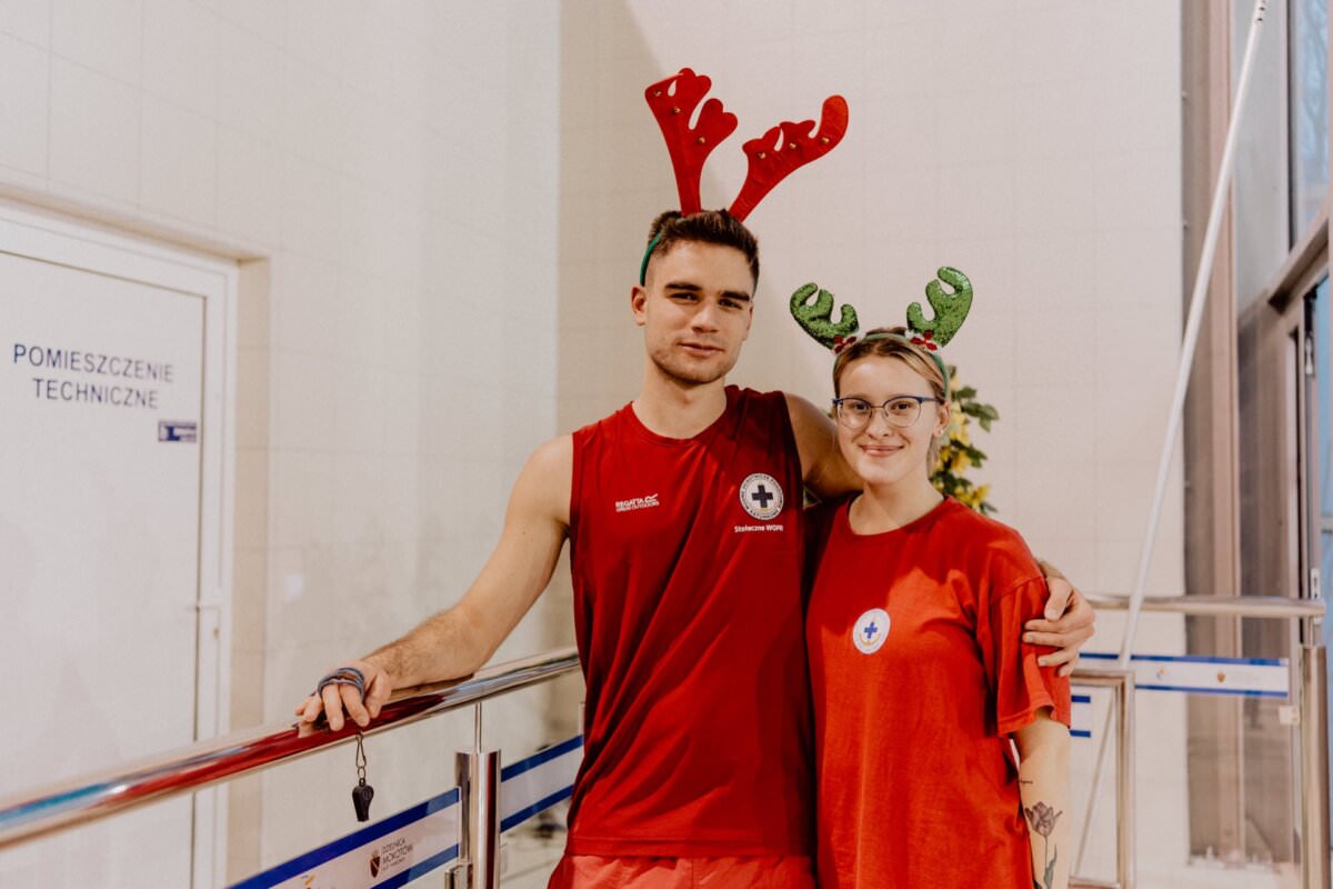 Dwie osoby ubrane w czerwone koszule i świąteczne poroże reniferów stoją obok siebie i uśmiechają się do kamery. Wydaje się, że znajdują się w pomieszczeniu zamkniętym, a na drzwiach za nimi widoczny jest napis „Położenie Techniczne”, uchwycony w ramach fotorelacji z wydarzenia zorganizowanej przez Marcina Krokowskiego.