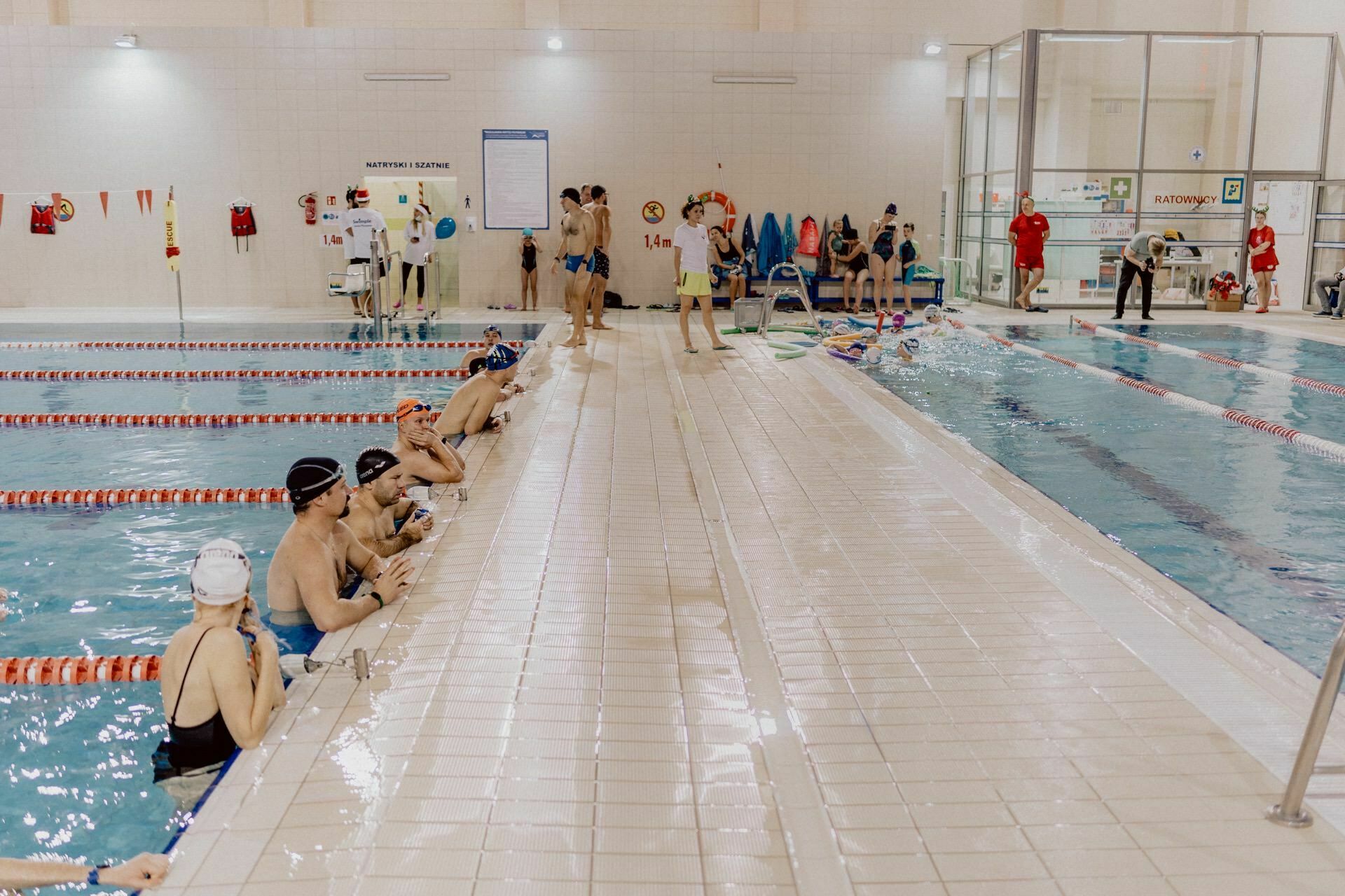 Swimmers wearing swimming caps and goggles relax at the edge of the indoor pool, where several lanes are set aside for practice. Coaches and other participants are present in the pool, engaging in activities and observing. The scene is captured by Marcin Krokowski, an acclaimed event photographer Warsaw.  