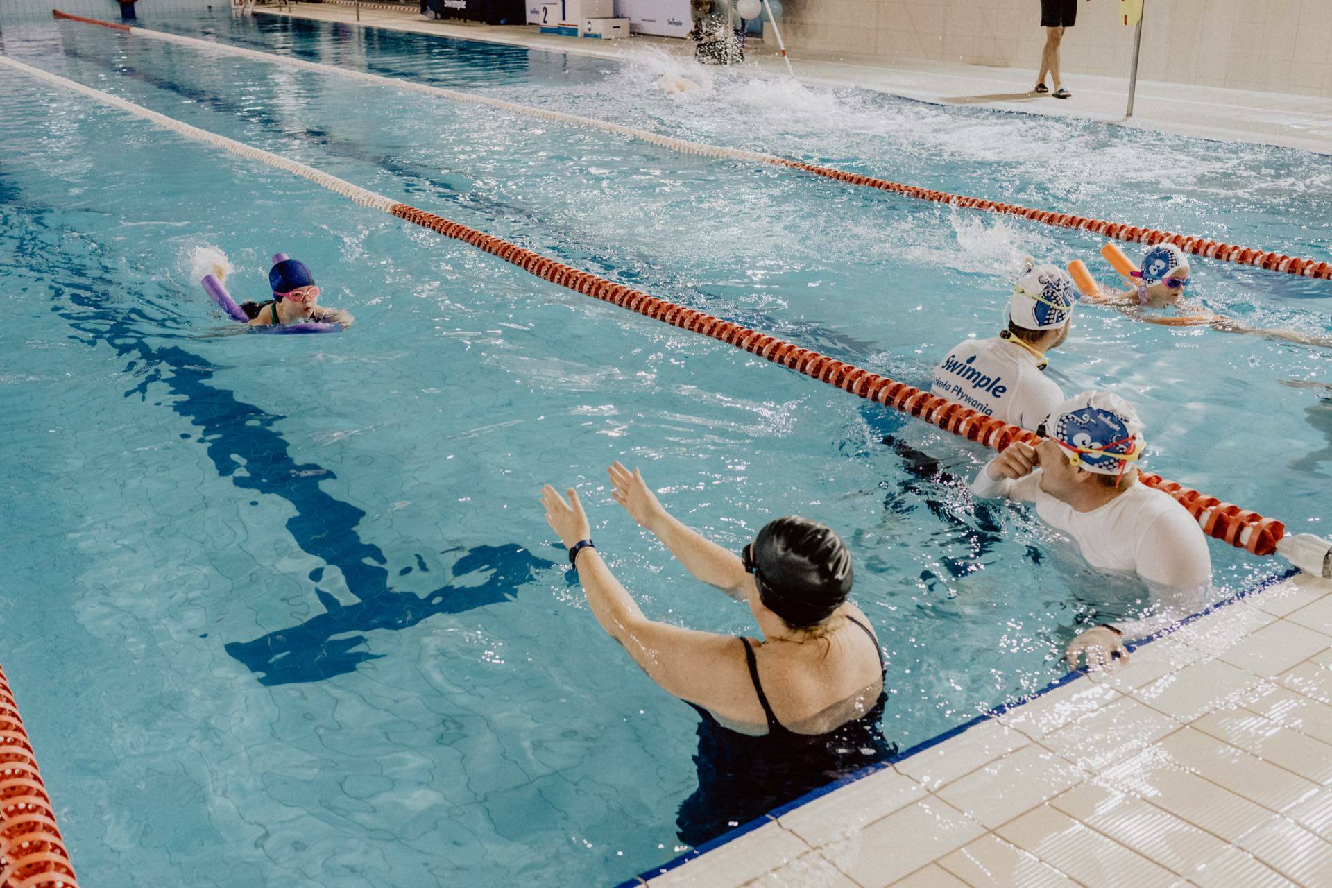 Several swimmers practicing in an indoor pool, separated by track dividers. A coach on the pool deck gives instructions while another coach and several swimmers are in the water, focusing on swimming techniques. Ideal for event photography that captures the energy and dedication of the athletes.  