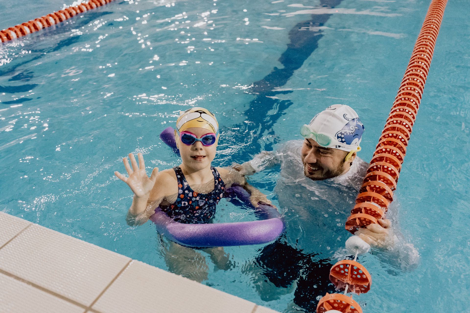 A young girl in a swimming cap and goggles waves while holding a purple pool noodle. An adult man in a swimming cap and goggles stands beside her, smiling and holding onto the railing separating the pool lanes. This may be part of an event photo essay by event photographer warsaw.  