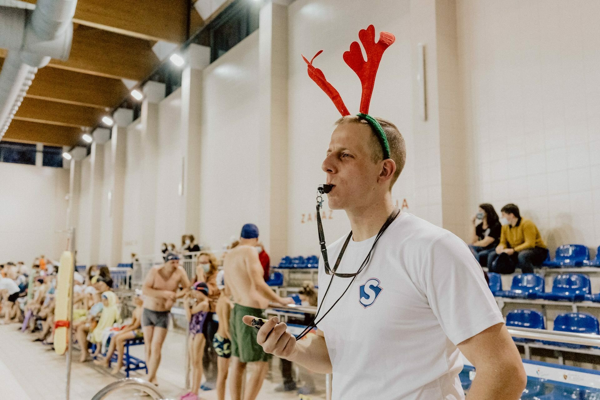 A swimming coach dressed in reindeer antlers and a white T-shirt with a logo uses a stopwatch and whistle to oversee the event. In the background are swimmers in swimsuits and people sitting in the stands, capturing dynamic photo coverage of the events. 