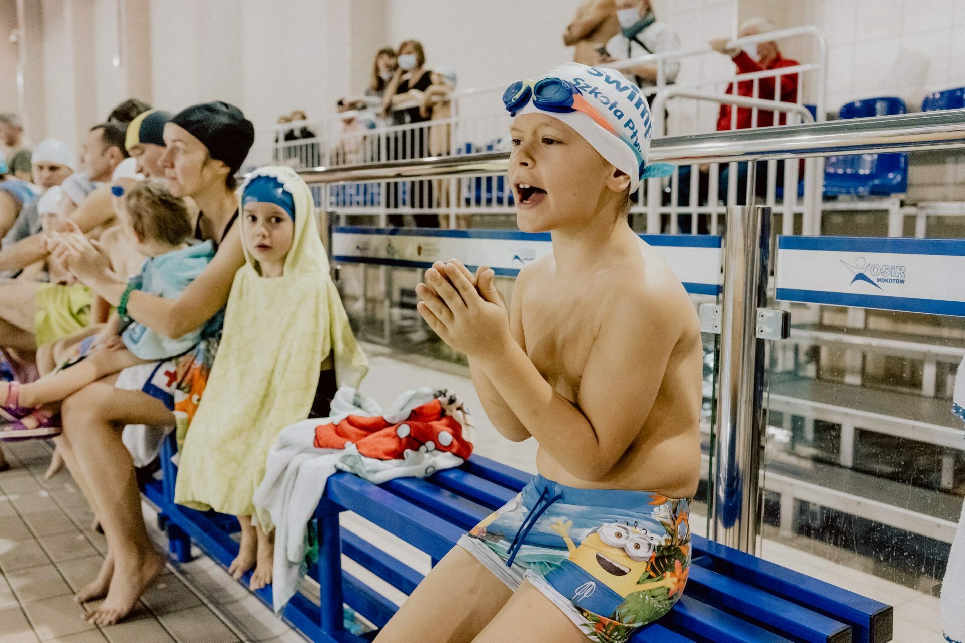 A young boy wearing swim trunks and a bathing cap with goggles squats on a bench and cheers enthusiastically. Nearby, a young girl wrapped in a yellow towel watches intently. Behind them, spectators sit on benches, some holding children. The bustling scene was perfectly captured by Marcin Krokowski, event photographer Warsaw.   