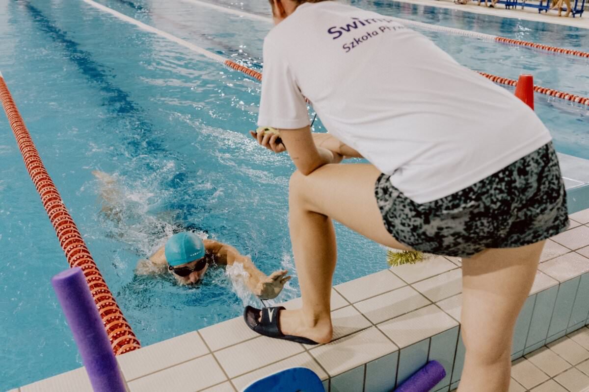 Pływak w niebieskiej czapce płynie torem na krytym basenie, podczas gdy osoba w białym t-shircie i czarnych spodenkach stoi na krawędzi, obserwując i prawdopodobnie mierząc czas pływaka. Na pierwszym planie widać wyposażenie basenu, takie jak makaron i płetwy, co uchwyciło autentyczny moment autorstwa fotografa eventowego Marcina Krokowskiego.