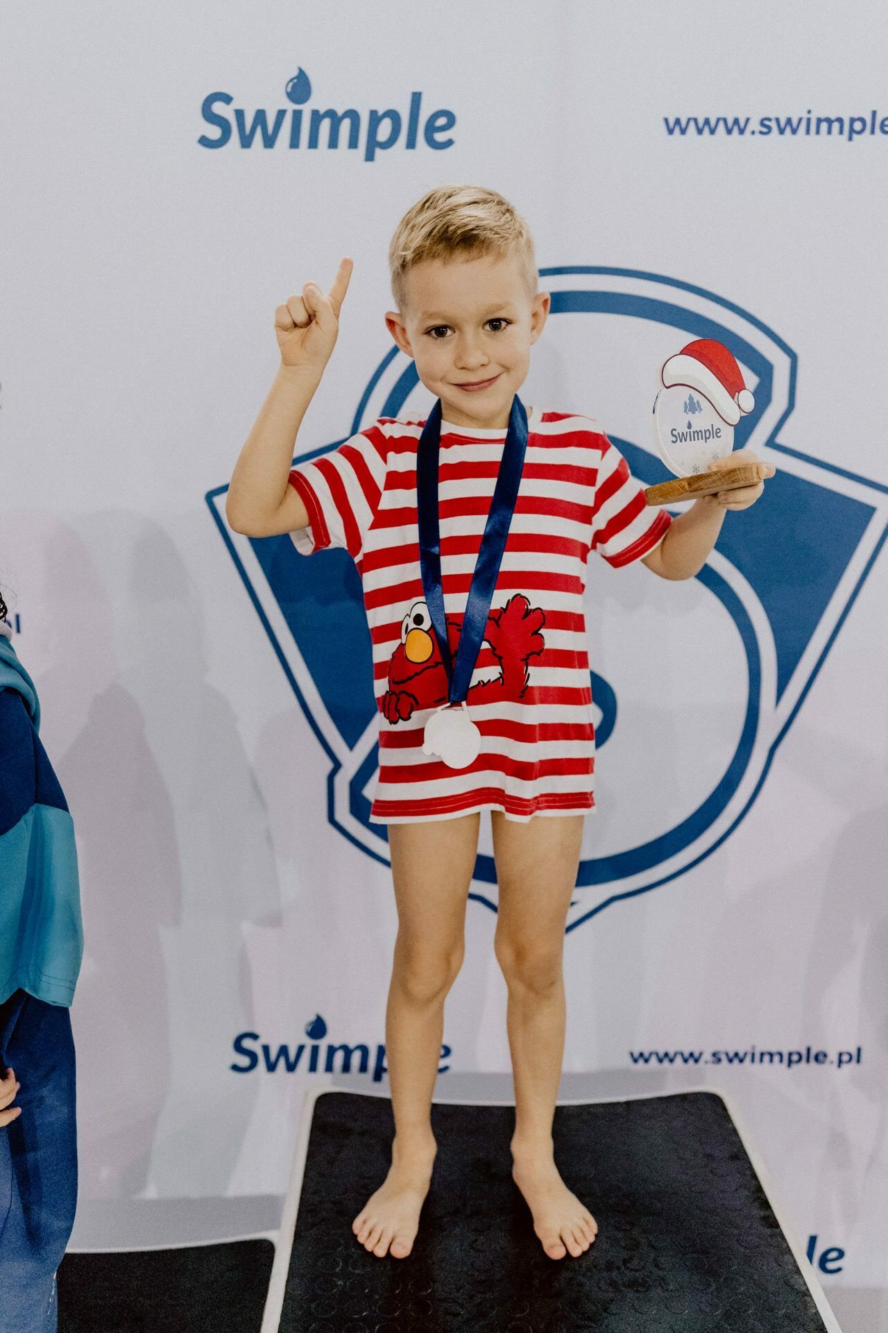 A young boy wearing a red-and-white-striped T-shirt, depicting a cartoon character, stands at a podium, holding a trophy. He wears the medal around his neck and smiles, pointing his finger upward. The logos "Swimple" and the website "www.swimple.pl" are visible in the background, captured perfectly by event photographer Marcin Krokowski.  