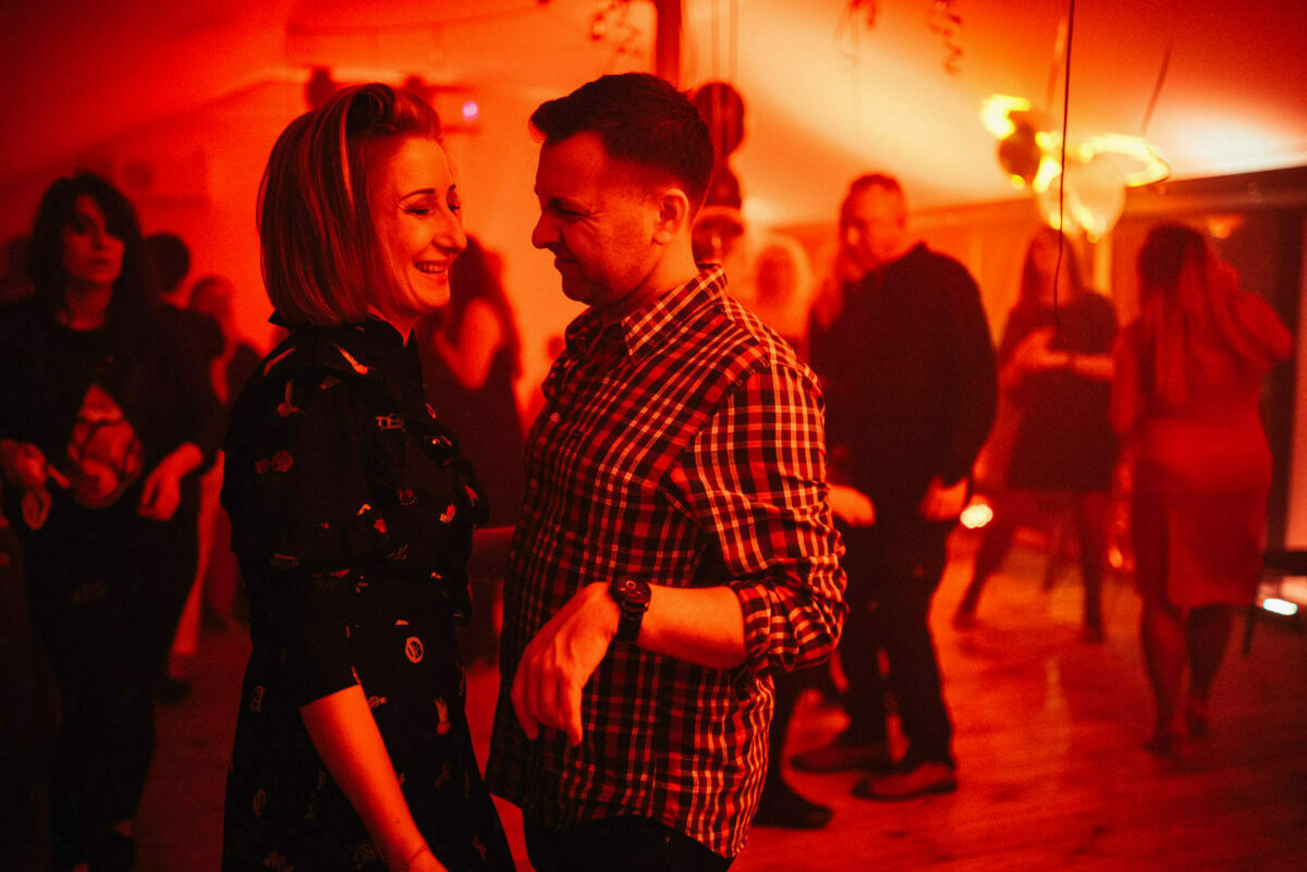 The couple is smiling and talking closely on a dimly lit dance floor illuminated by red light, surrounded by other people dancing and talking. The atmosphere is warm and festive, suggesting a party or social gathering captured beautifully in the event photo report. 