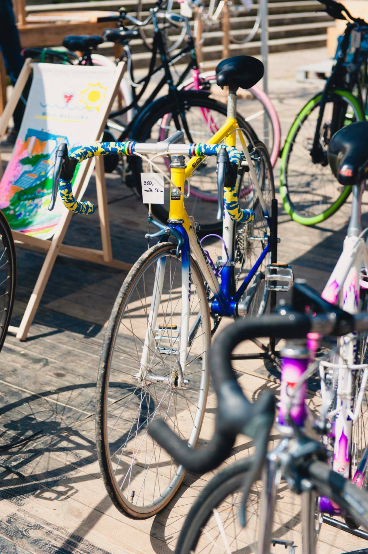 A colorful collection of bicycles parked outside on a sunny day. One of them stands out with a yellow and blue frame with a striped handlebar tape. In the background is a deckchair with a bright, abstract pattern and a €250 price tag - the perfect shot for any event photographer capturing photo coverage of the fair.  