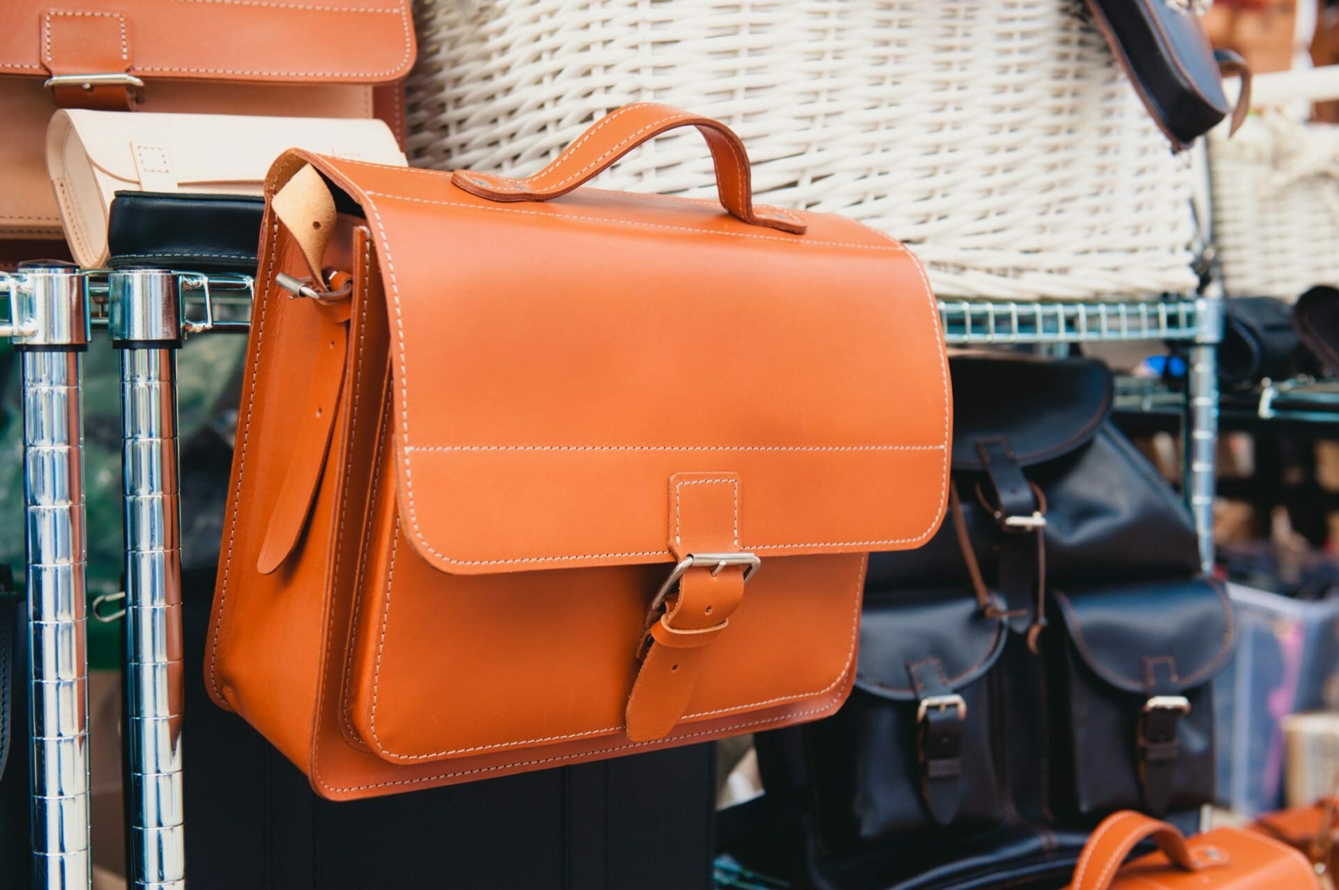 A brown leather handbag with a buckle strap is displayed on a rack, beautifully captured by the event photographer. All around are other bags, including black leather bags and cream wicker baskets in the background, all neatly arranged and displayed on shelves - a perfect photo-op. 