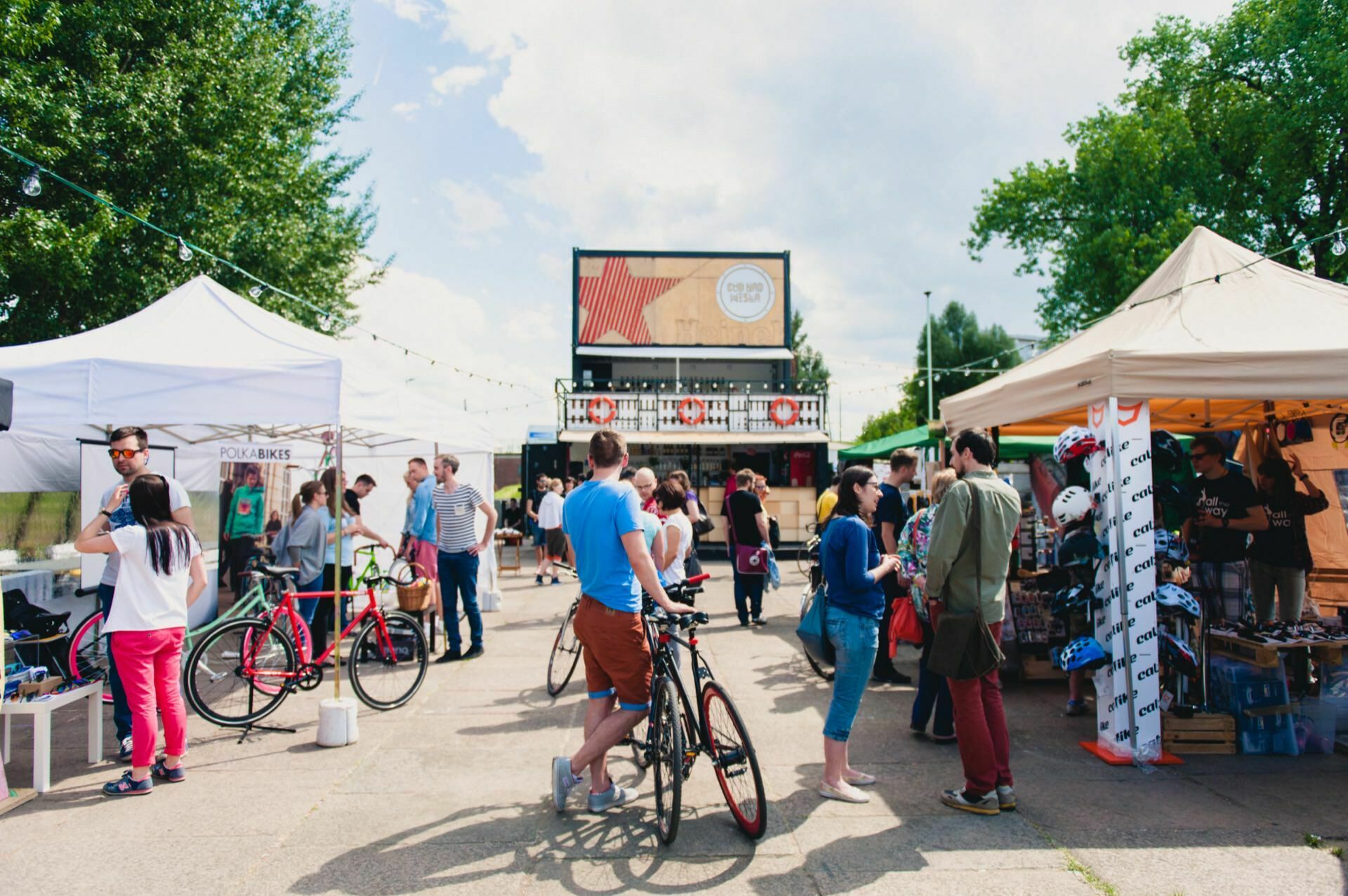 A bustling open-air market where people shop, talk and walk around. Various stalls and tents sell goods, including a bicycle store. Some hold bicycles or ride them. In the background you can see trees and a billboard with a sign - the perfect setting for a photo report of the event by a photographer from Warsaw.   