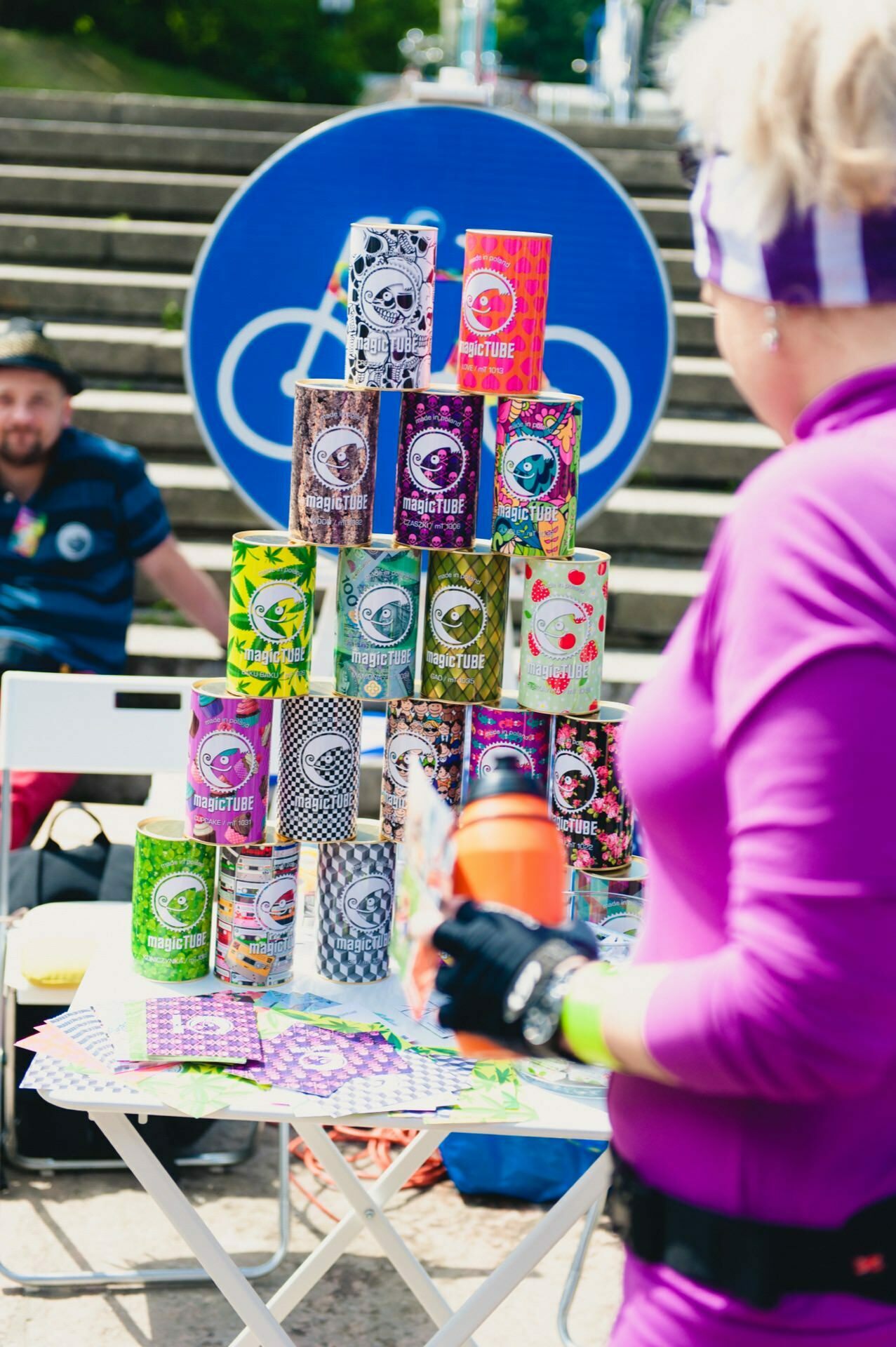 A person in purple attire is standing in front of a table with a pyramid of colorful cylindrical cans of various designs. In the background, a man sits on a chair by a large blue sign with a white bicycle icon. The scene looks outdoors, which appears to be a photo-op of a trade show.  