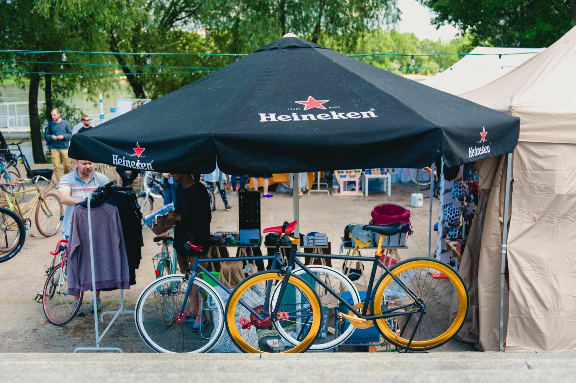 An outdoor market scene with a black Heineken umbrella shading a tent filled with various items for sale. Several bicycles are parked in front of the building, including a distinctive yellow and black one. Shoppers browse while trees are in the background, creating the perfect setting for a photo essay of the fair.  