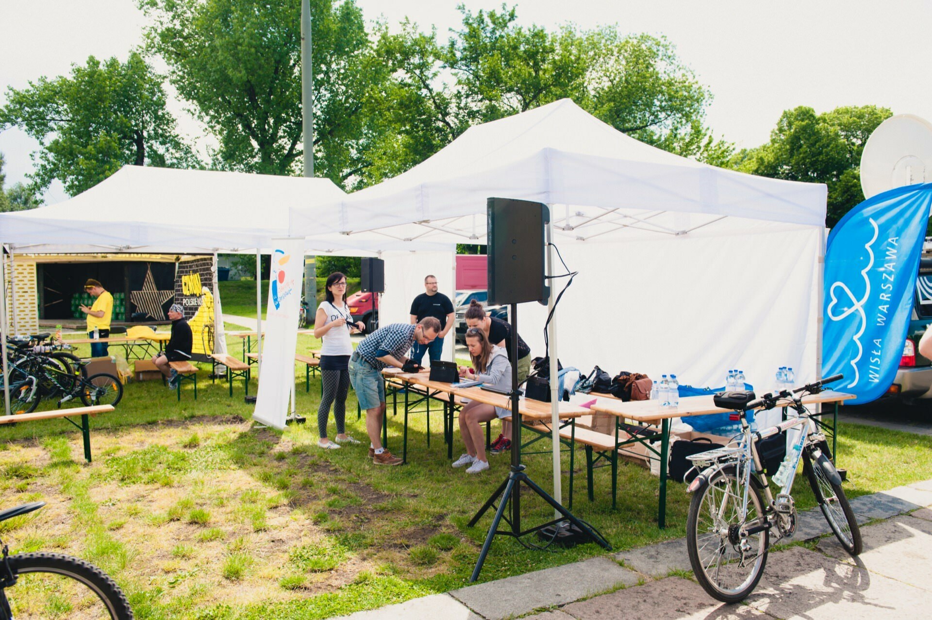 An outdoor event with people gathered under white tents on a sunny day. A few people sit at a table with bottles of water and papers, others stand nearby. Bicycles stand on the grass. In the background is a blue banner with white text and heart symbols, capturing the essence of an event photographer.   