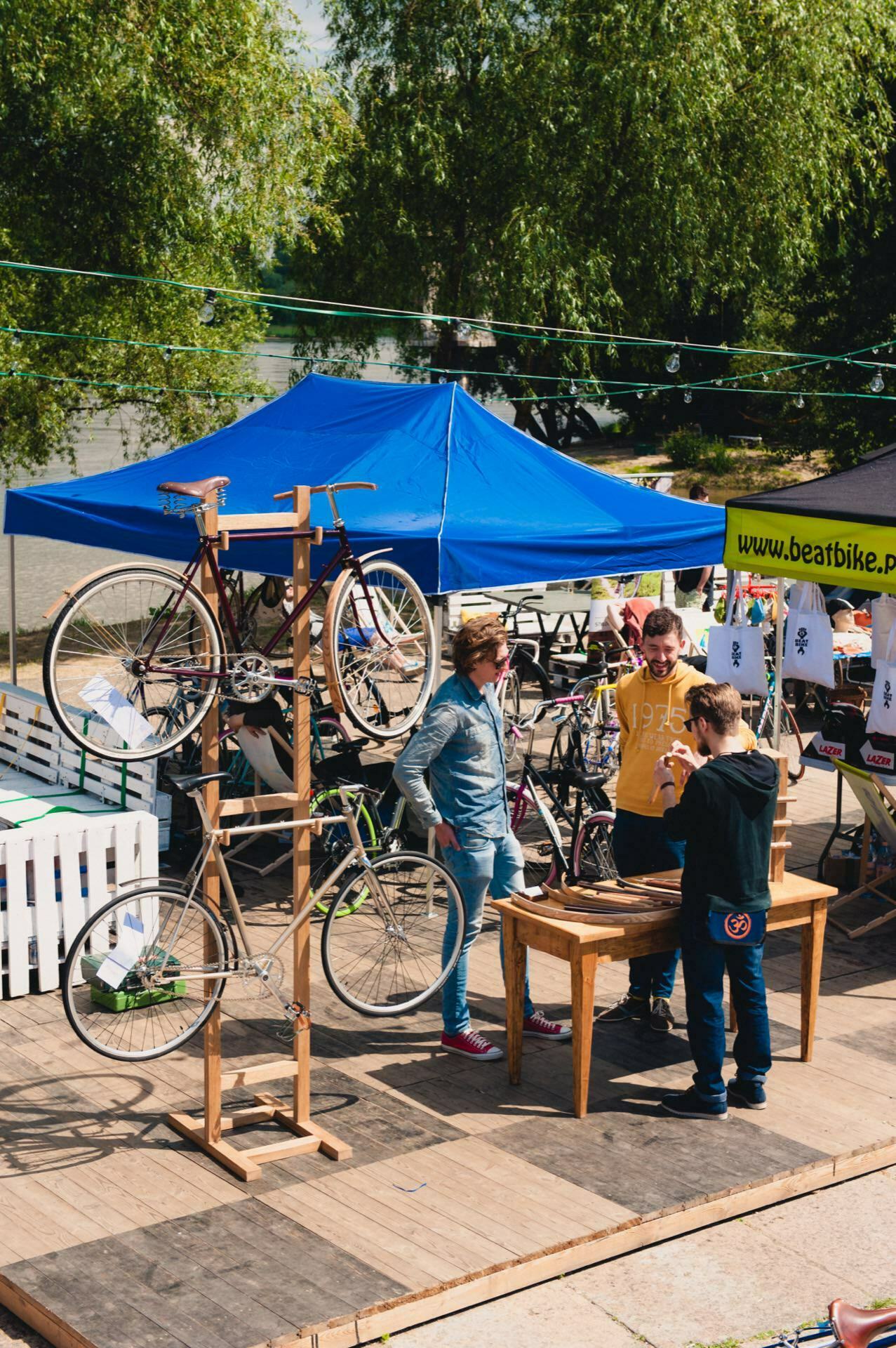 The centerpiece of this vibrant event is an outdoor bike repair station with a blue tent and several bicycles on display. An event photographer captures the moment as three people gather around a wooden table immersed in conversation. Trees and the river create a picturesque backdrop with strings of lights overhead, and a white fence and banner complete this charming photo story of the fair.  