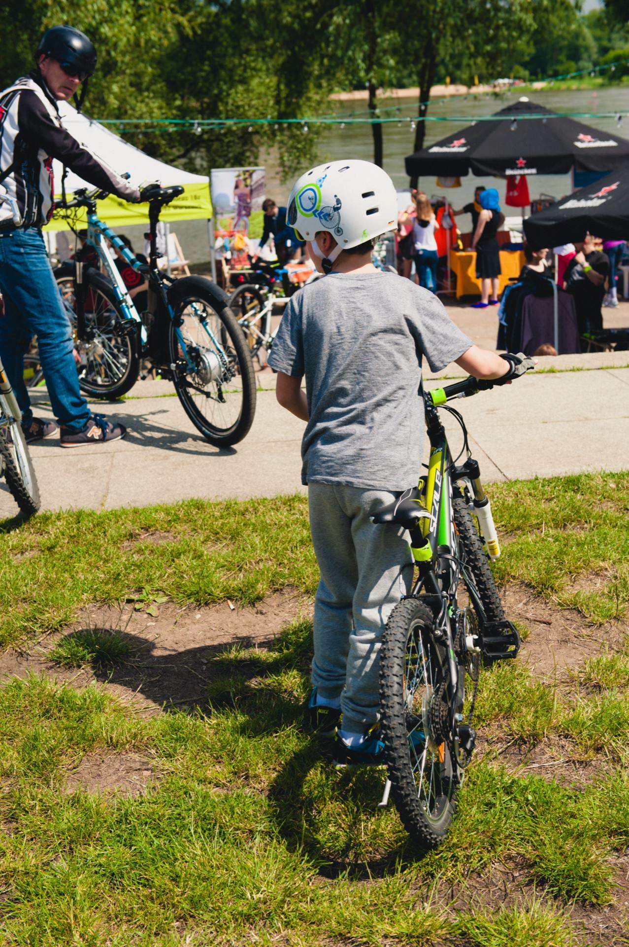 A child in a white helmet and gray clothing stands next to a bicycle on a grassy area. An adult in bicycle gear adjusts the bike nearby, capturing the essence of the scene like an event photographer. In the background, people gather under tents and trees in this park-like setting, as if it were part of a photo tour of the fair.  