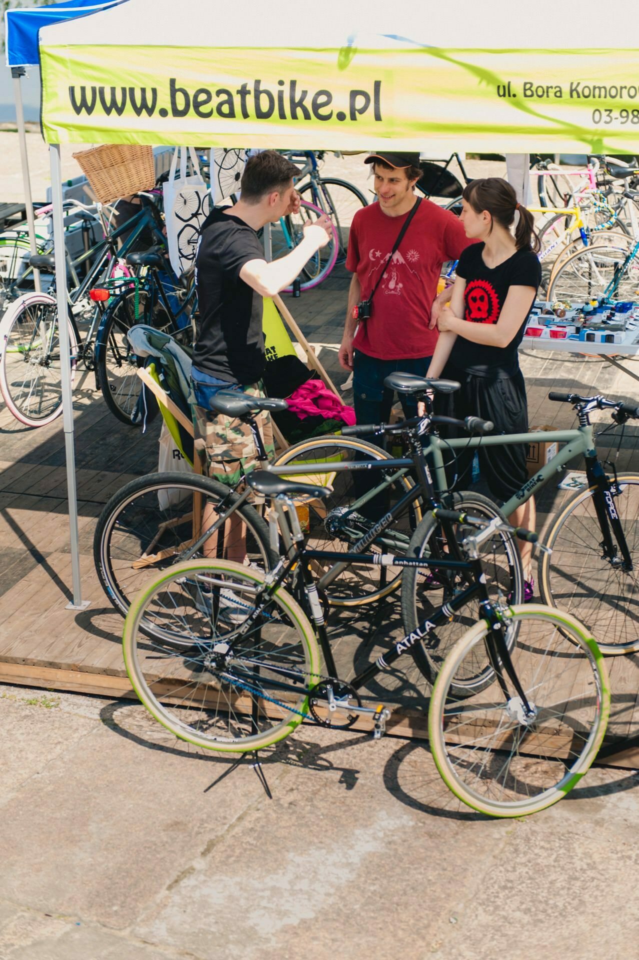 Three people are standing under a yellow canopy tent with the URL "www.beatbike.pl" on it. One person, surrounded by several bicycles, gestures, while another holds a tool. This scene, ideal for an event photographer, seems to capture a lively discussion or demonstration related to cycling.  