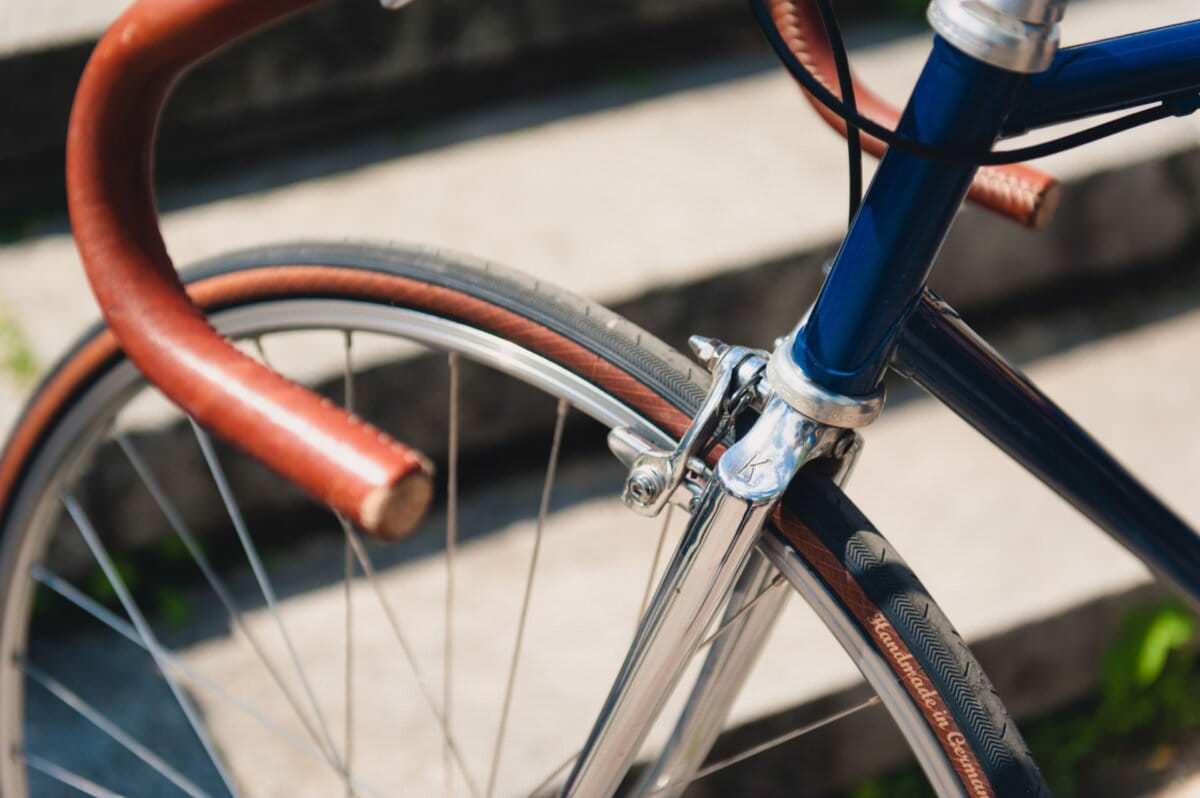 Zbliżenie na przednie koło niebieskiego roweru zabytkowego, przedstawiające obszytą skórą kierownicę i srebrny przedni widelec. W tle znajdują się schody z szarego kamienia, a światło słoneczne rzuca na scenę pstrokate cienie – doskonale uchwycając ten moment, tak jak fotograf eventowy zrobiłby to w fotorelacji z targów.